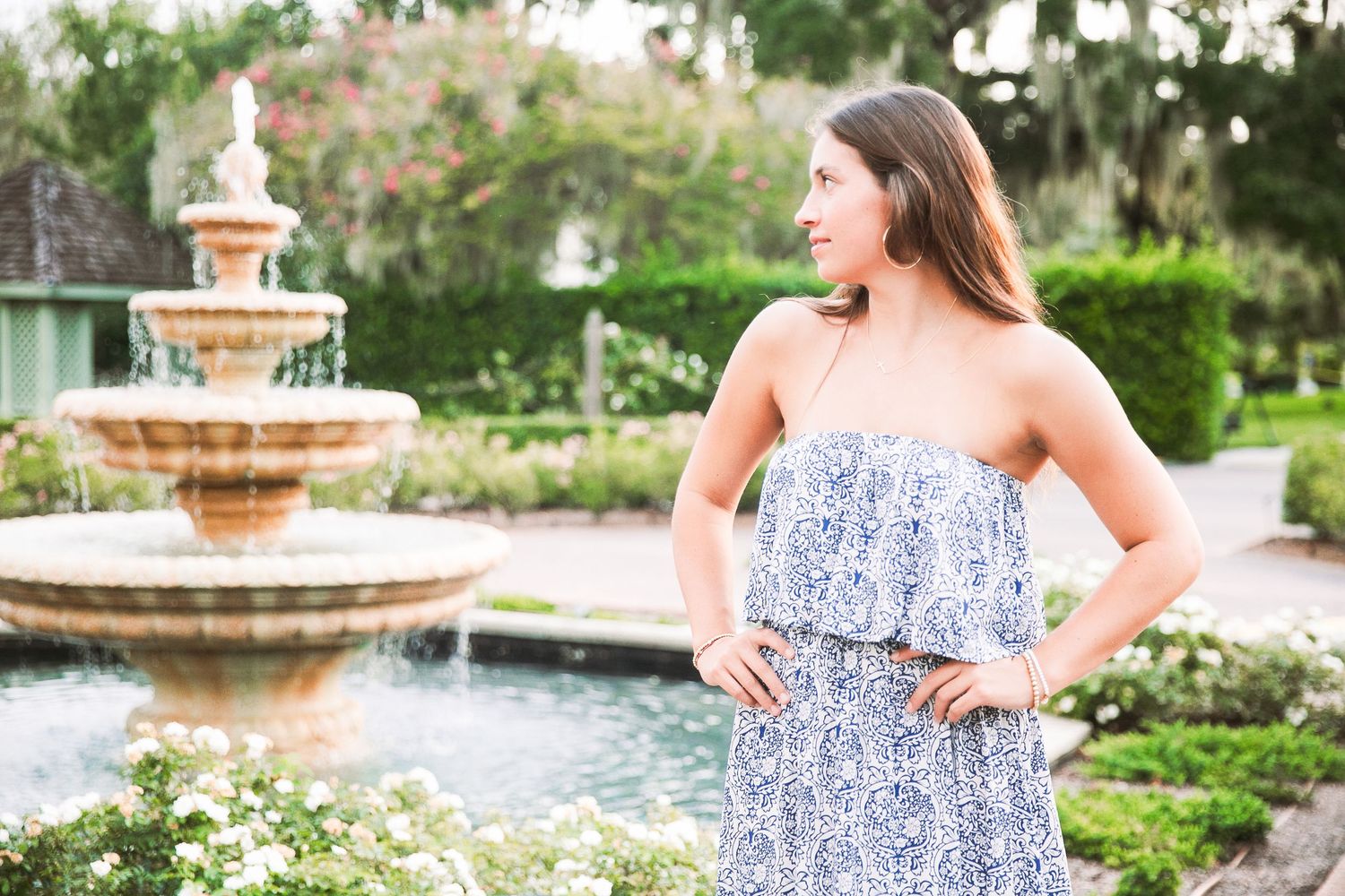 A person in a blue and white patterned sundress poses by an elegant tiered fountain in a lush garden setting.