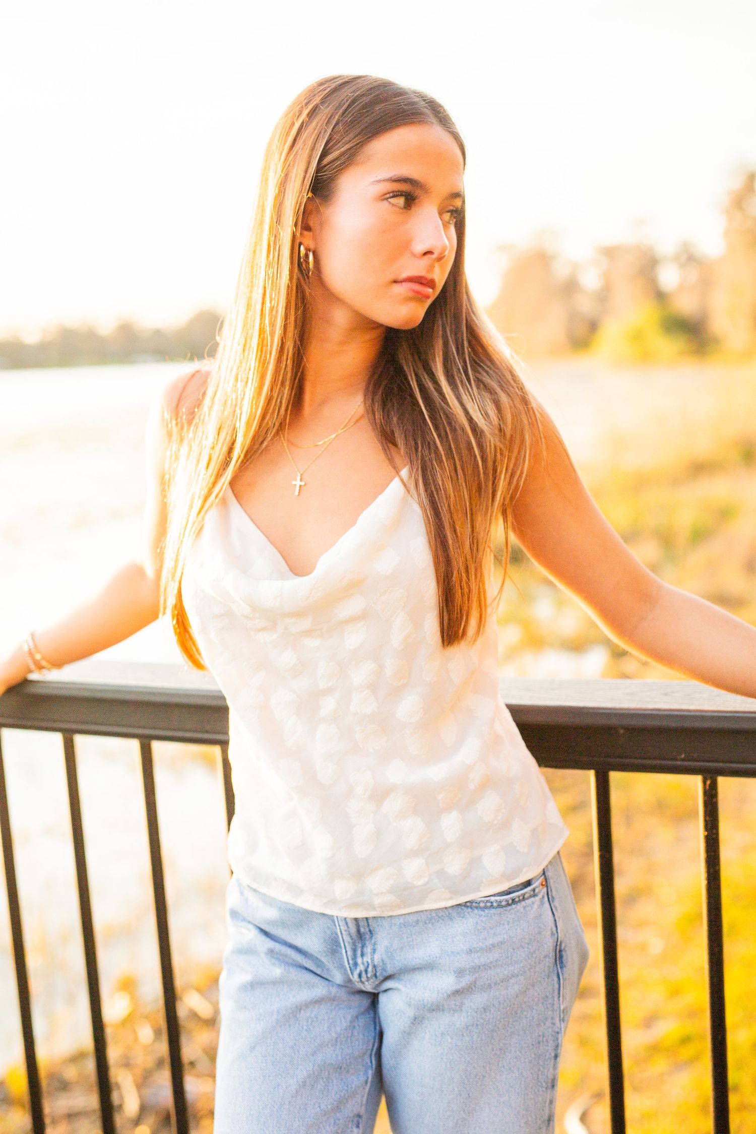 Person in white tank top and jeans standing by railing at golden sunset in outdoor setting.