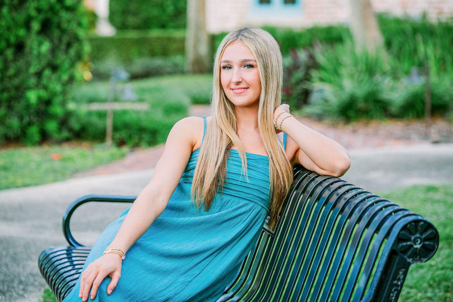 Person in turquoise dress sits relaxed on black metal park bench surrounded by lush garden landscaping.