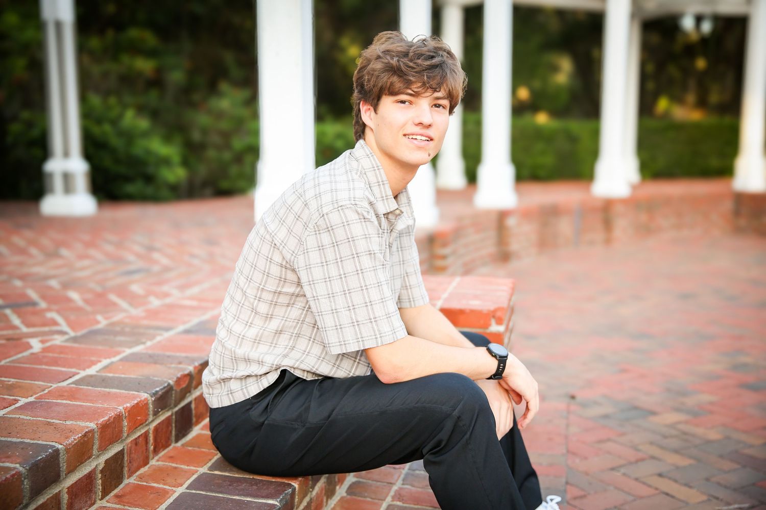 Someone sits casually on brick steps wearing a plaid button-down shirt near white pillars in an outdoor setting.