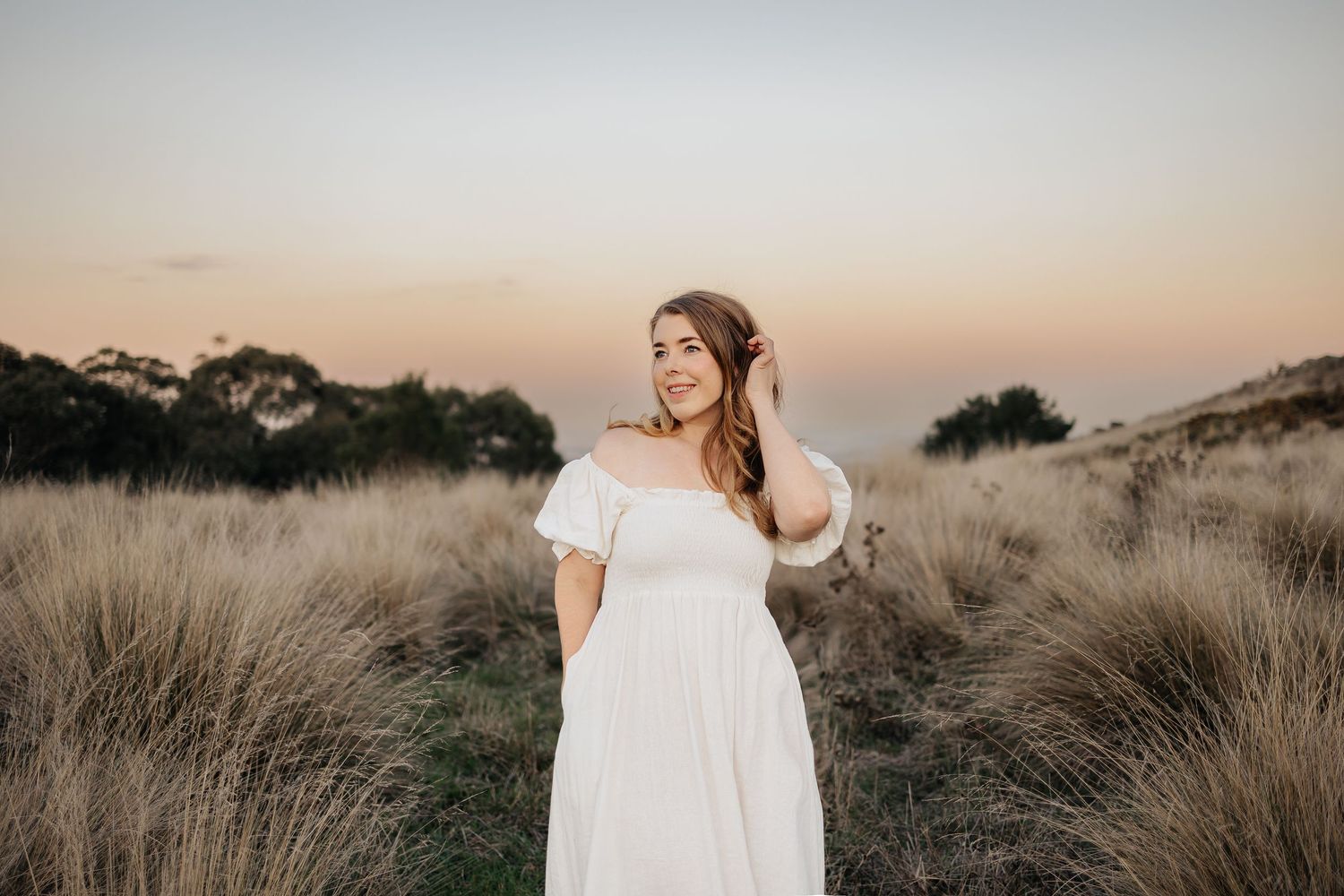 A woman in a white dress stands amid tall coastal grass during golden hour sunset.