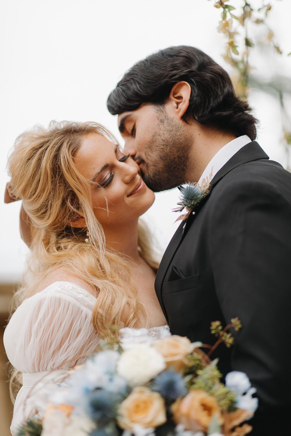 A tender wedding moment captured as newlyweds share an intimate kiss while holding a bouquet of peach and blue flowers.