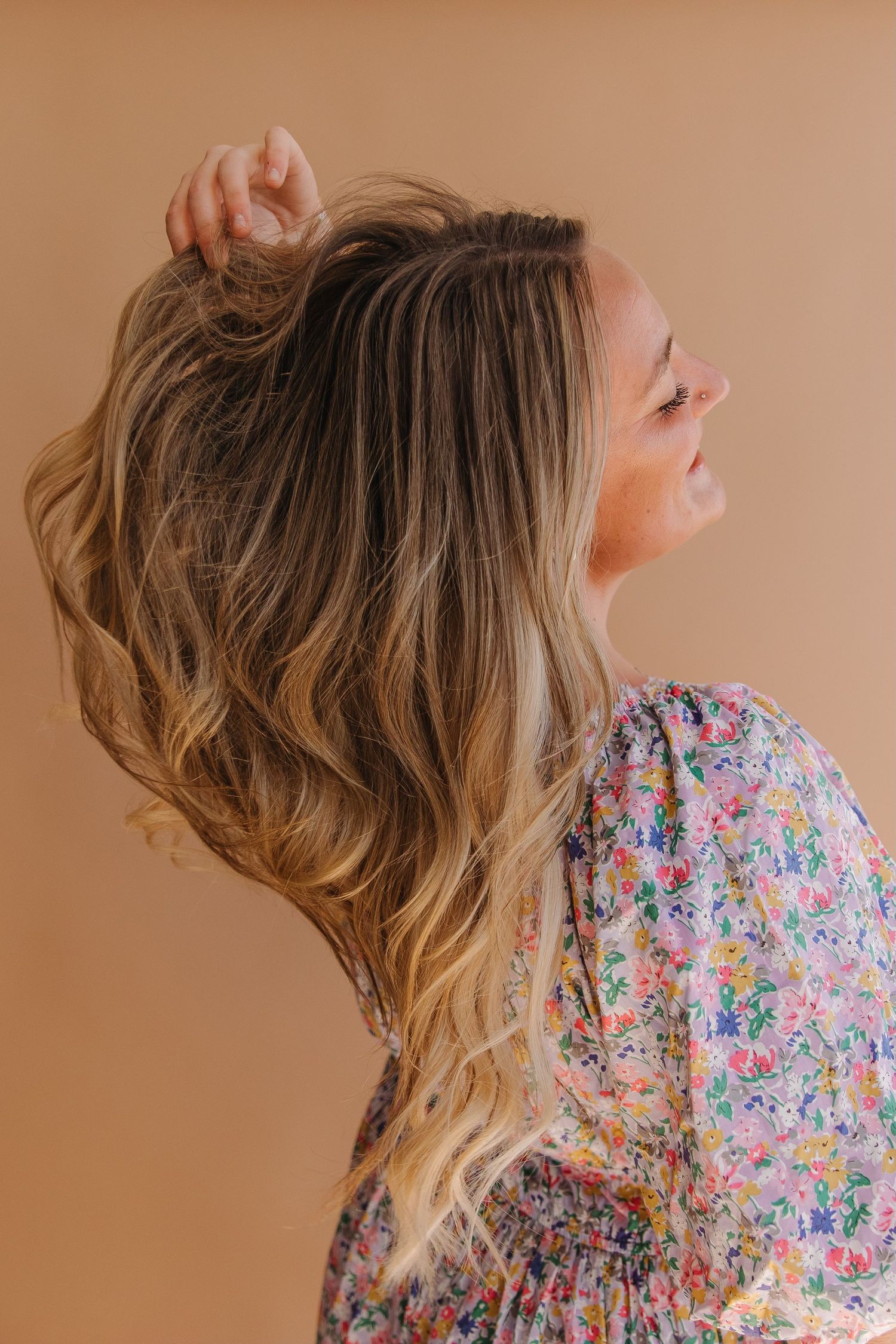 Woman with wavy blonde hair wearing a floral dress poses against peachy brown background in profile view.
