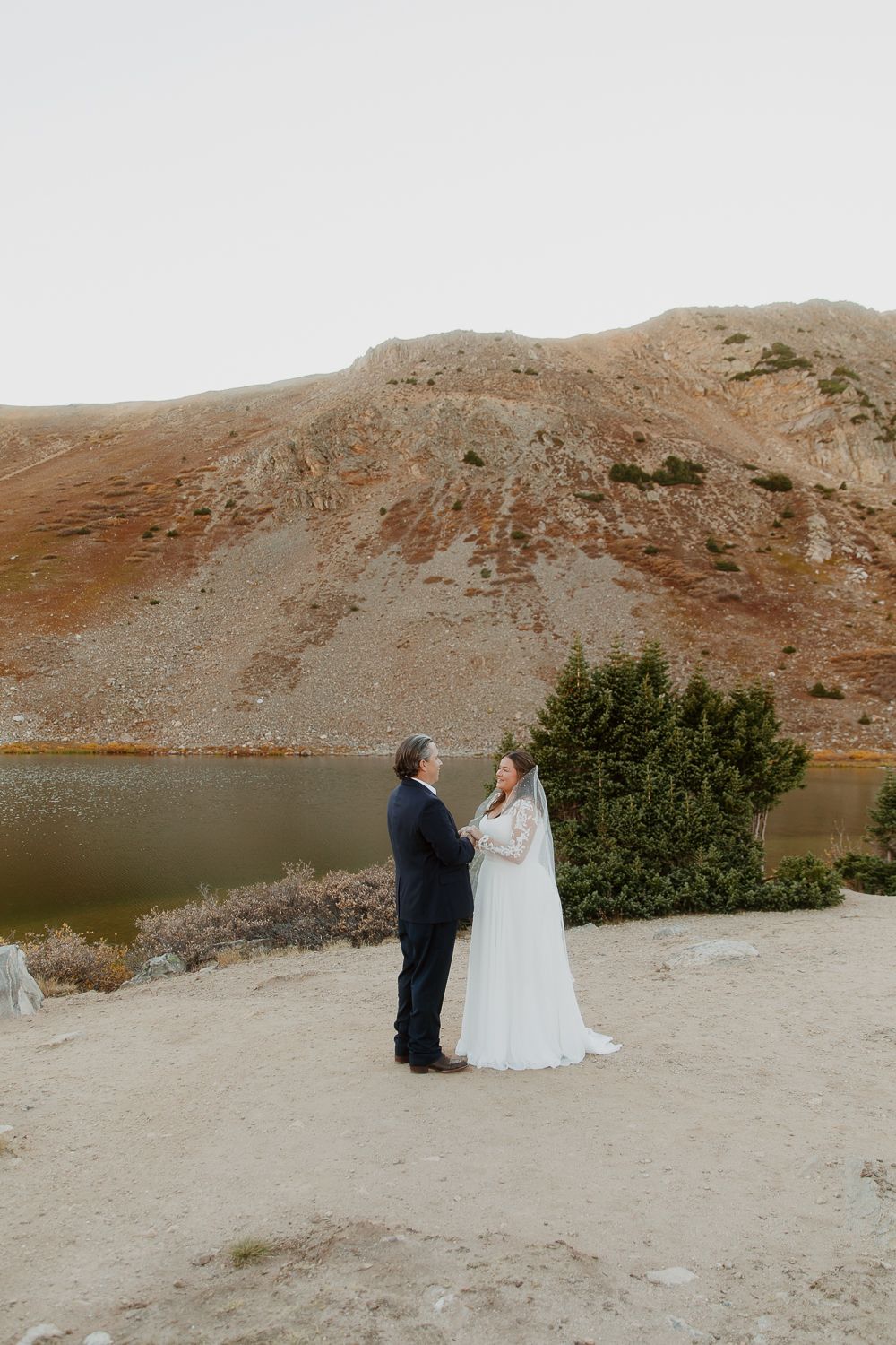 A couple in wedding attire stands together against a scenic backdrop of reddish mountains and a serene lake.