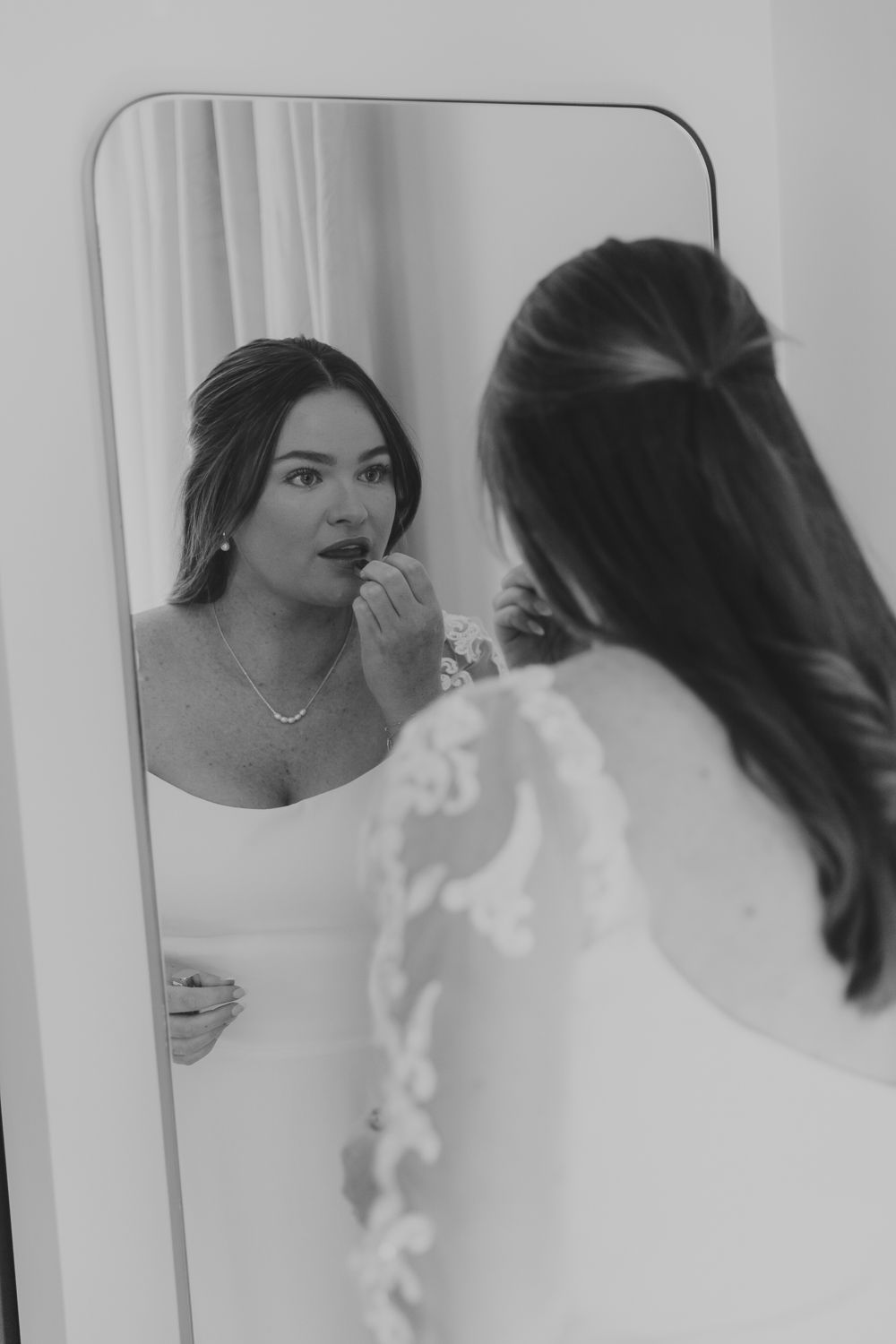 A person applies lip color while looking in a mirror, captured in elegant black and white photography.