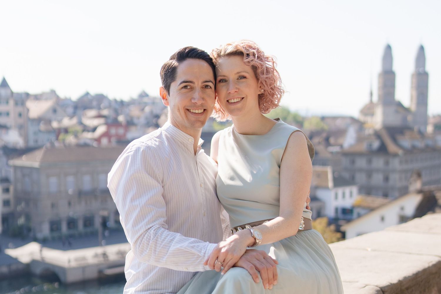 A couple in vintage inspired clothing embrace while sitting on a rooftop overlooking a historic European cityscape.