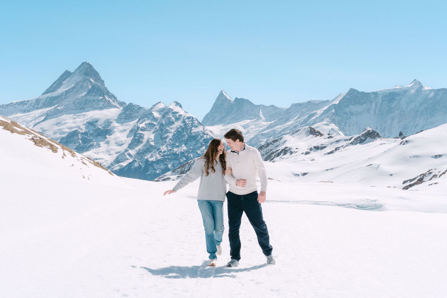 Two people embrace while standing in snowy mountains with dramatic alpine peaks in background against blue sky.