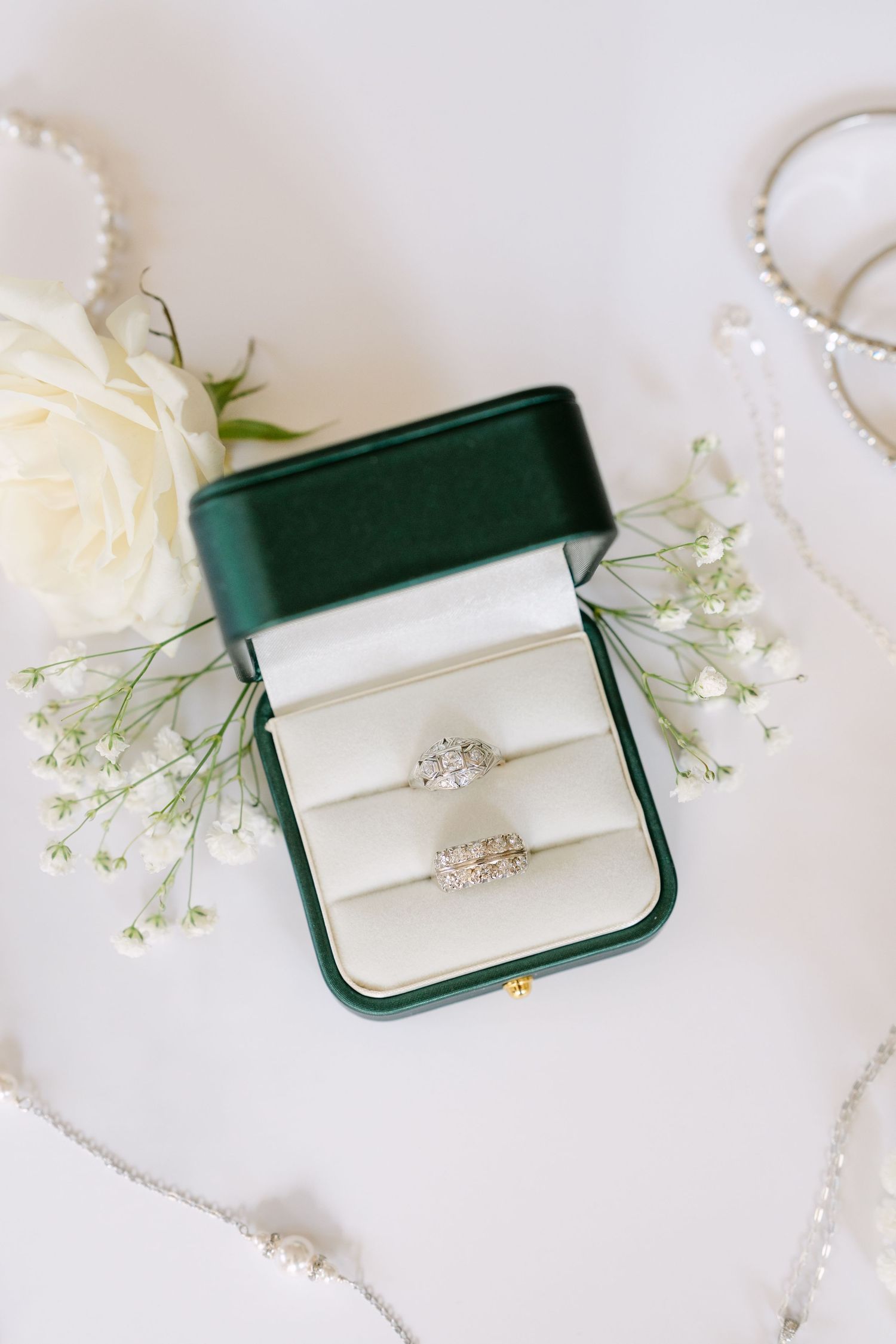 Two diamond engagement rings displayed in green velvet box with white rose and baby's breath flowers on white background.