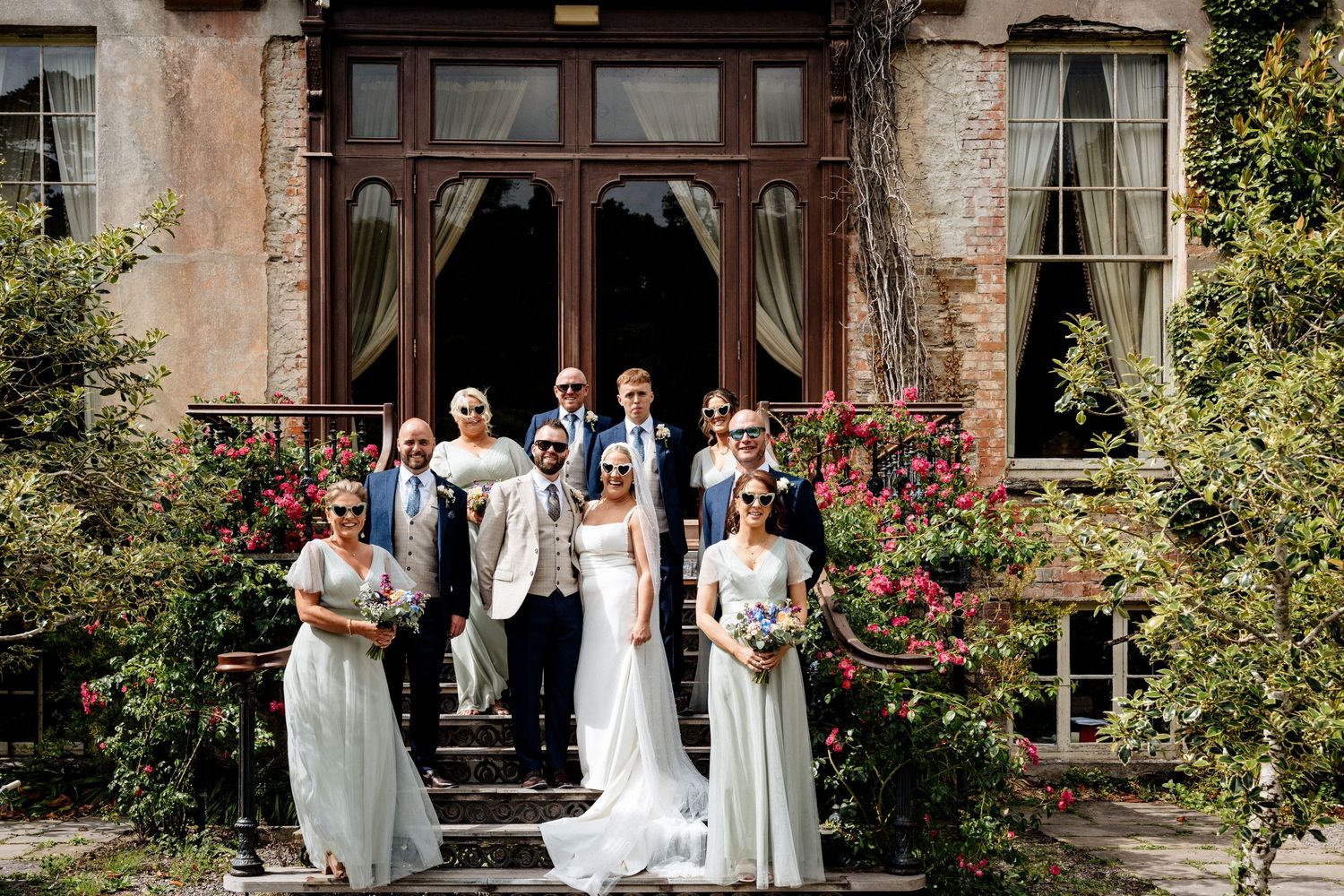 Wedding party poses on the steps of an elegant stone manor house adorned with climbing roses in full bloom.