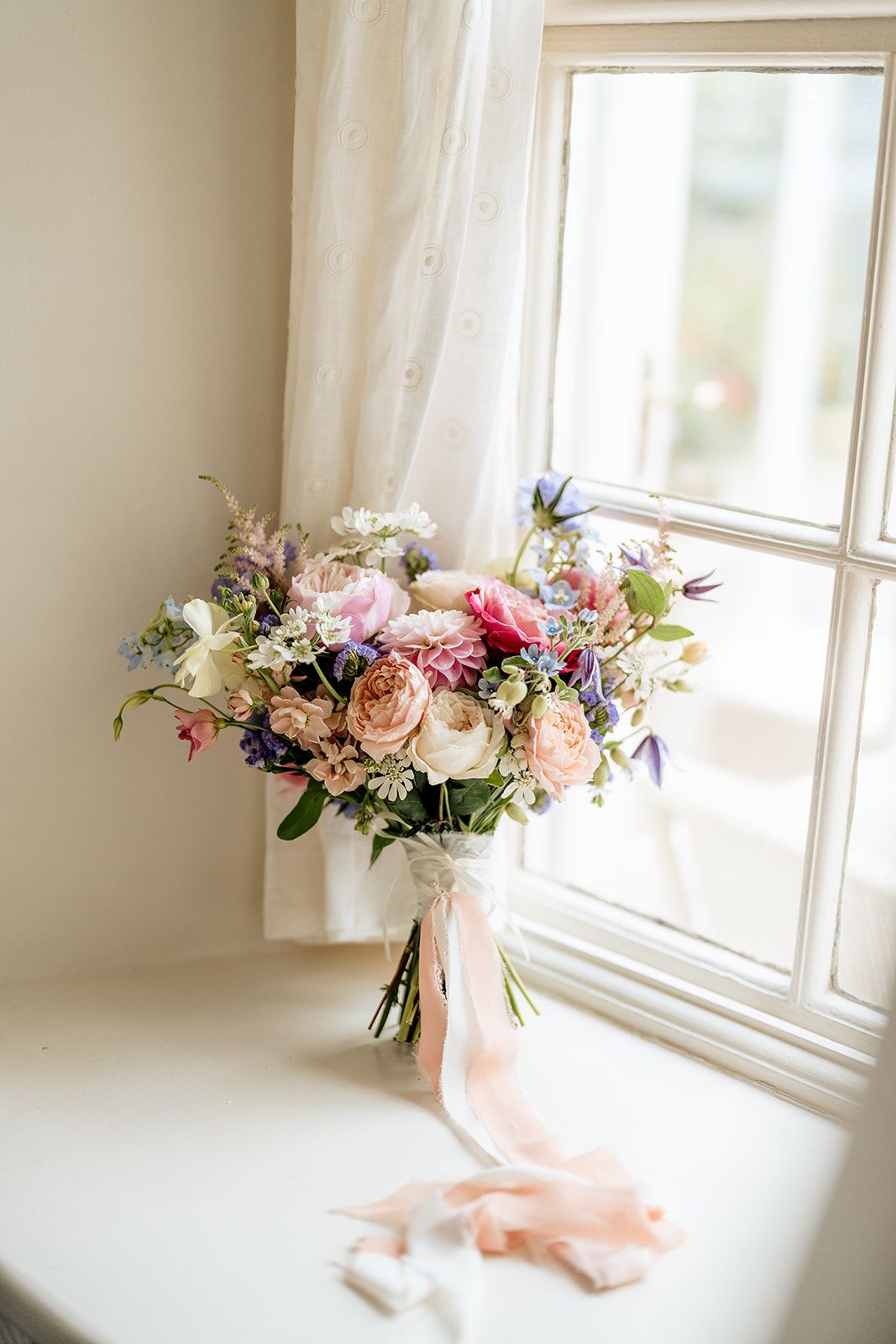 Beautiful wedding bouquet with pink and peach roses, wildflowers and trailing silk ribbons sitting by a sunny window.
