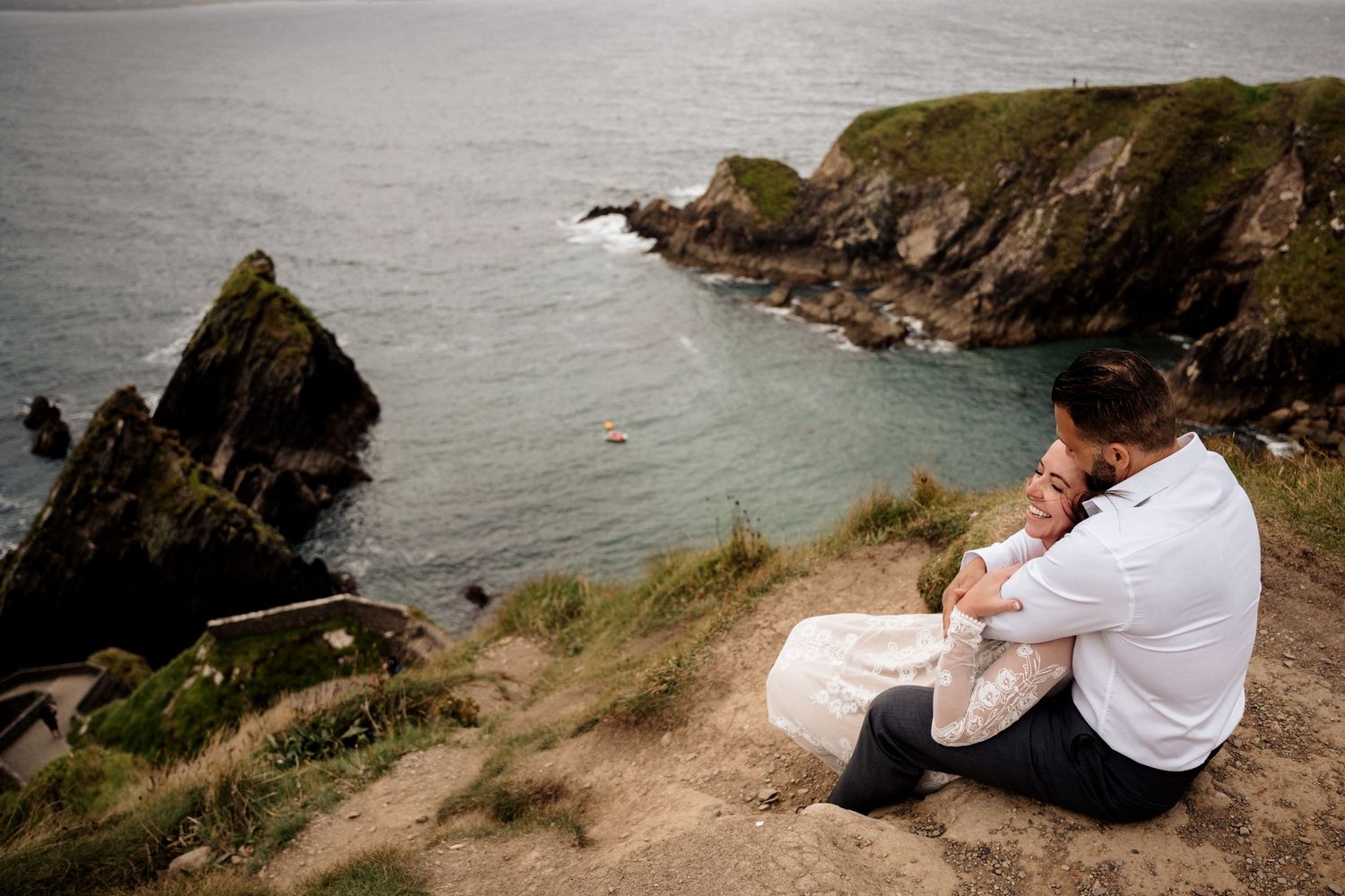 A couple rests on a cliff overlooking a dramatic rocky coastline with ocean views under moody skies.