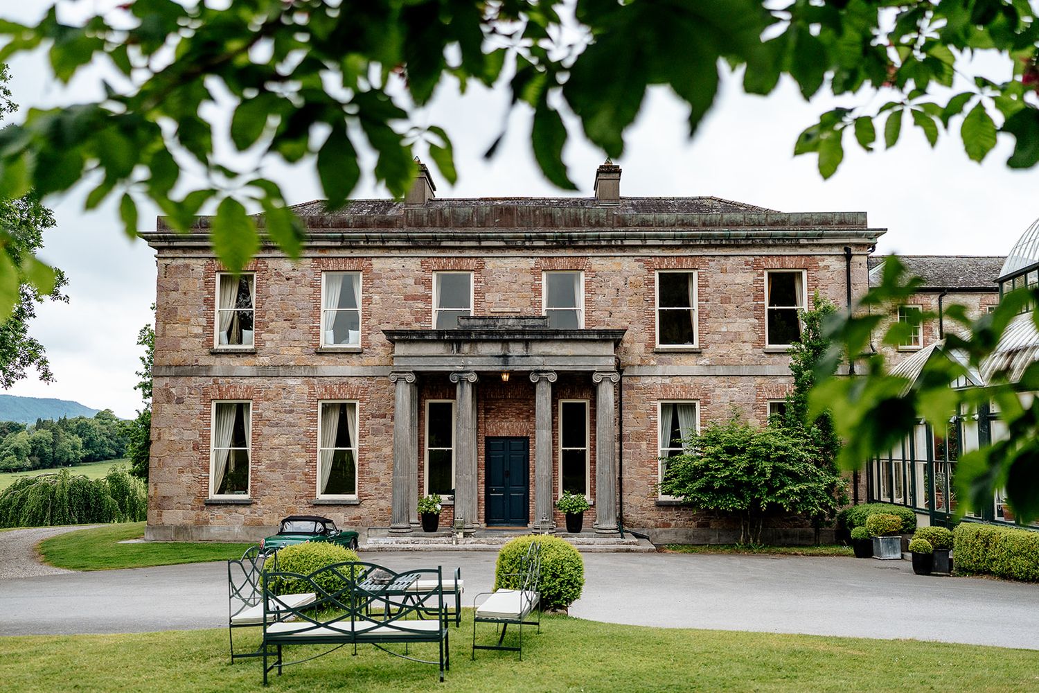 Historic Georgian mansion with red brick facade and columned entrance framed by green foliage and outdoor seating area.