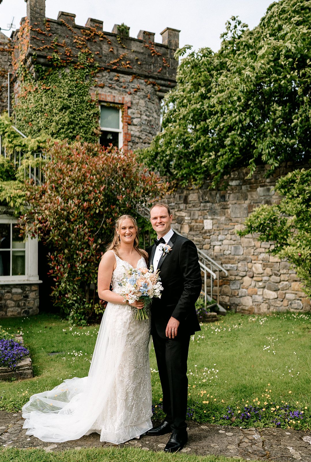Bride and groom pose in a garden beside stone castle wall during their wedding celebration.