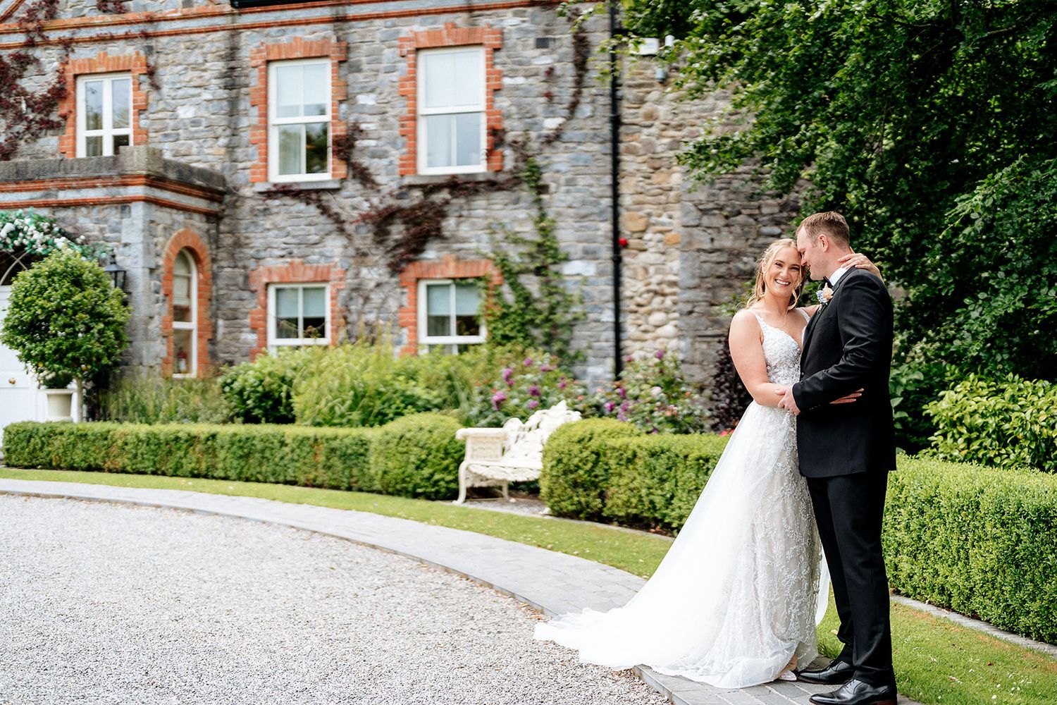 Romantic wedding couple embraces in front of stone mansion with manicured hedges and classic architecture.