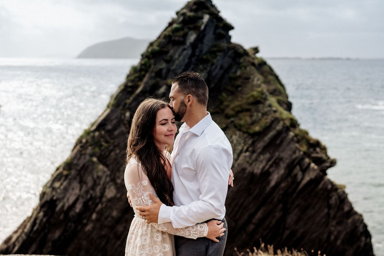 Two people share an intimate moment against a backdrop of dramatic coastal rock formations and misty sea.