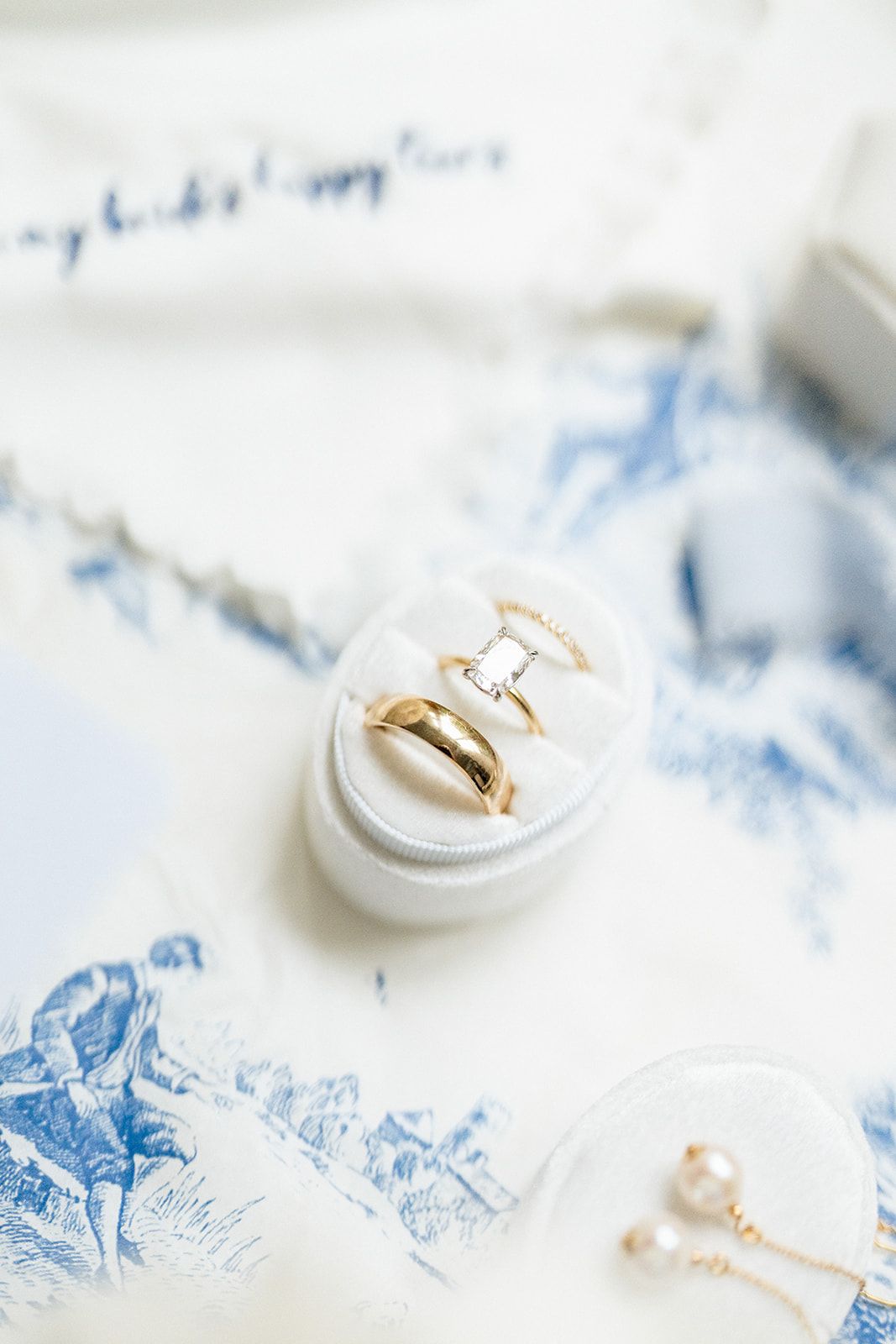 White jewelry ring box holding delicate gold rings on a blue and white patterned textile background.