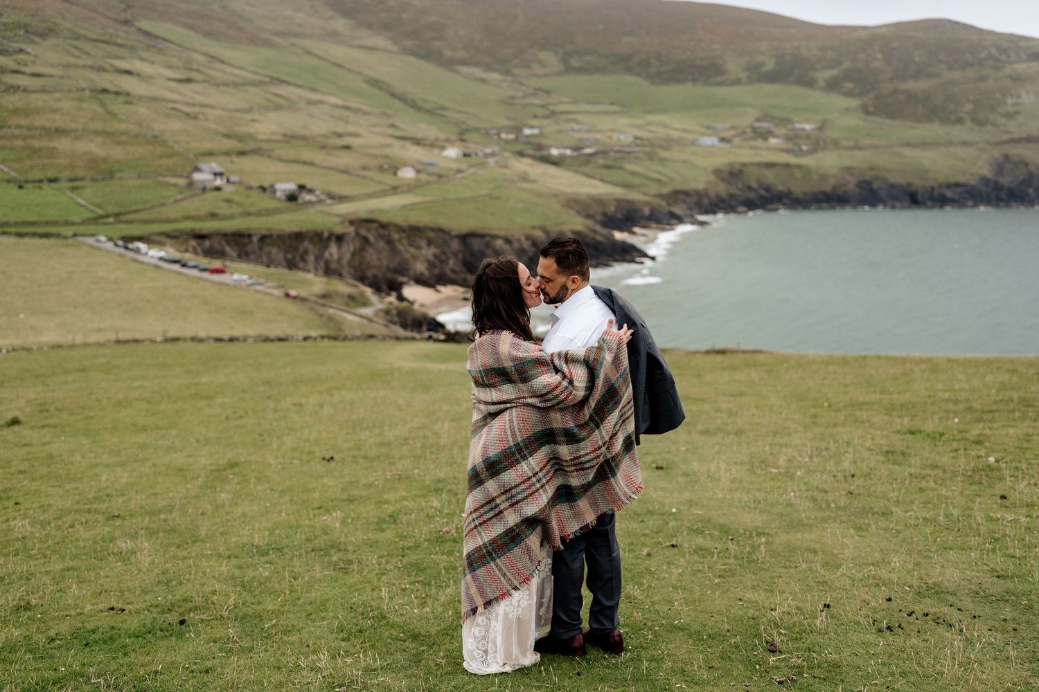 A romantic moment unfolds on green coastal cliffs overlooking a scenic bay with rolling hills.