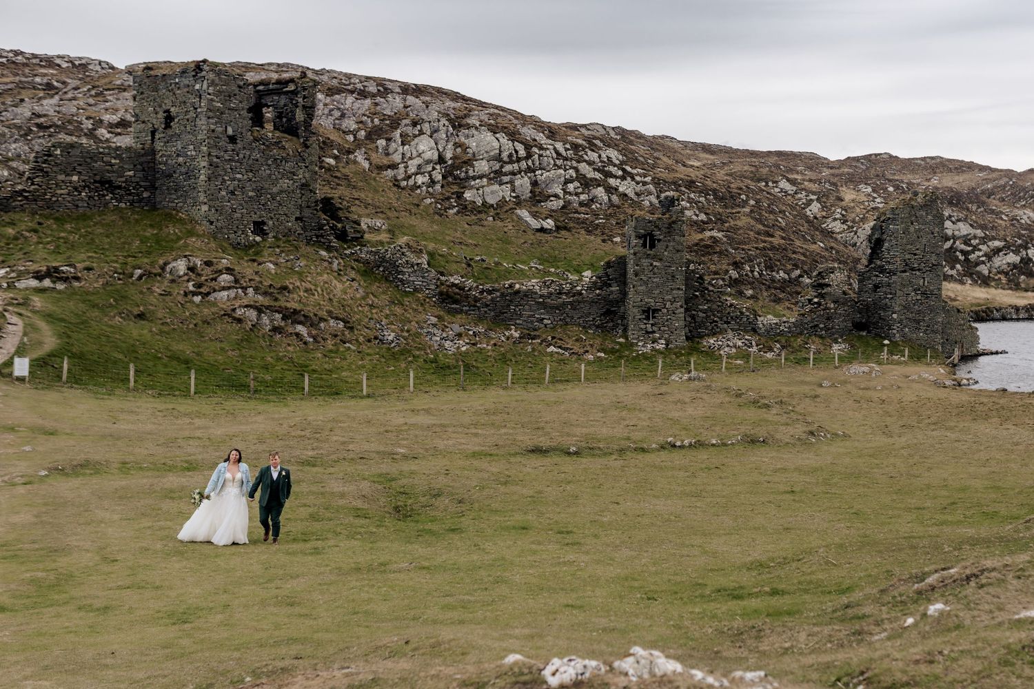 Couple walking near ancient stone ruins on a rugged Scottish hillside overlooking water.