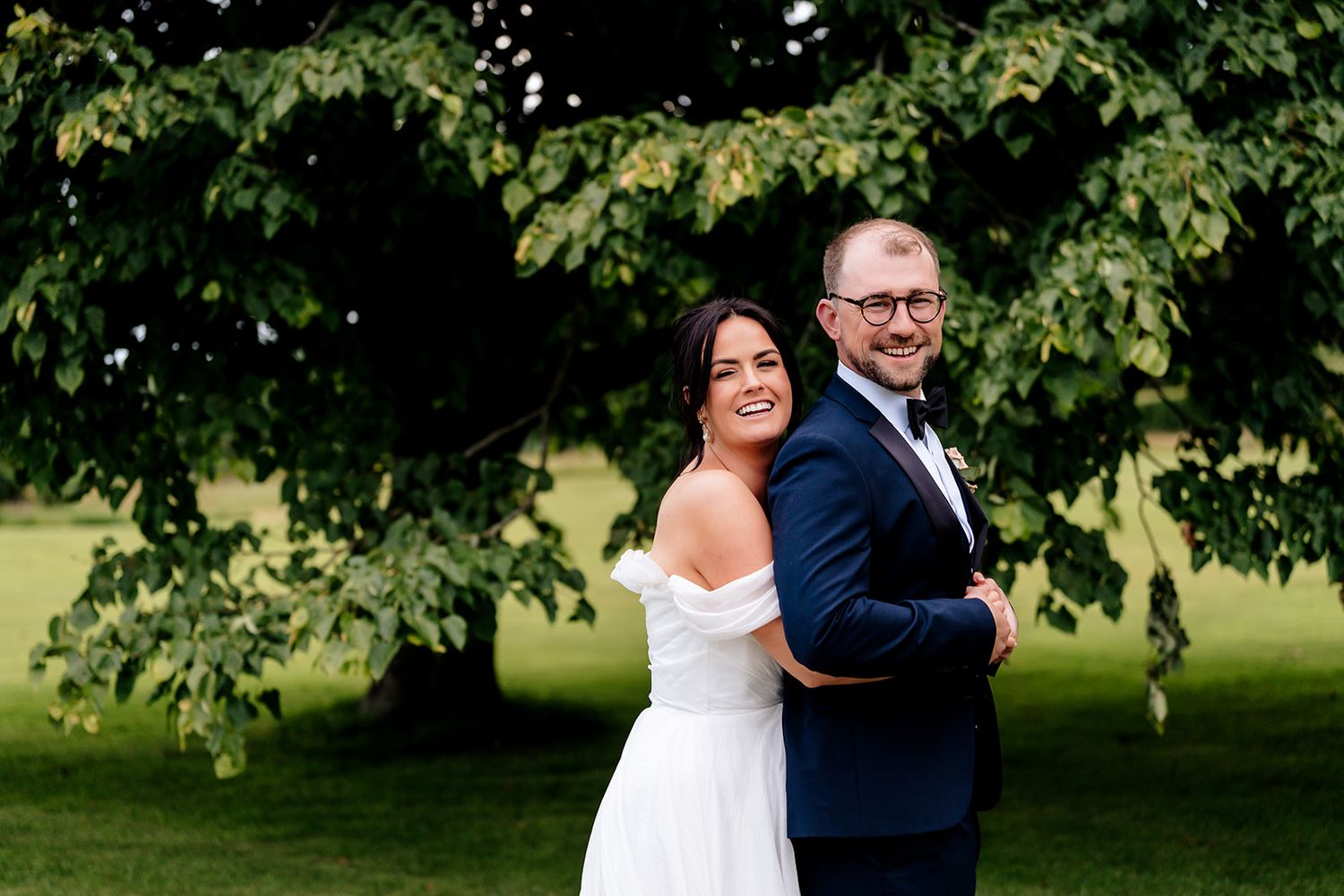 A couple in formal wedding attire pose back to back under a leafy green tree in a garden setting.