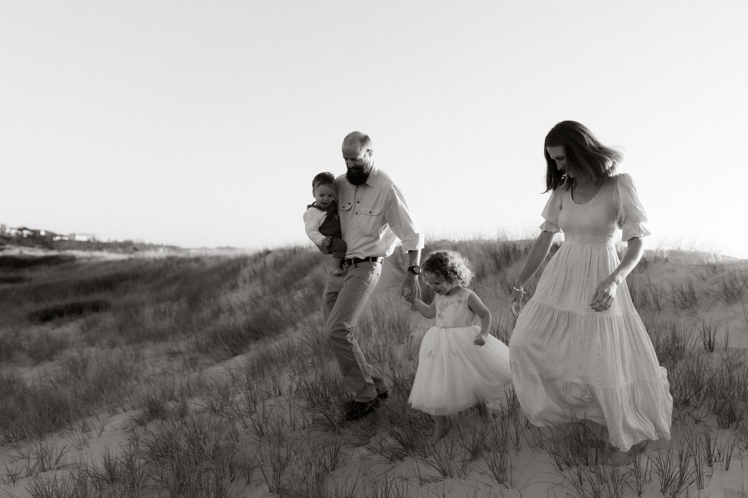 Family running down a grassy sand dune together at sunrise, black and white