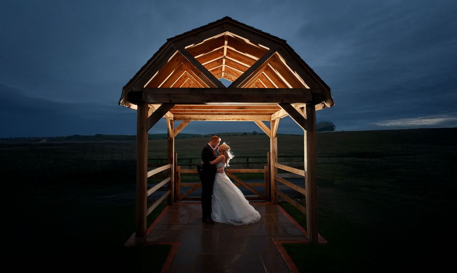 A couple embraces under a warmly lit wooden gazebo at dusk with dramatic stormy skies in the background.