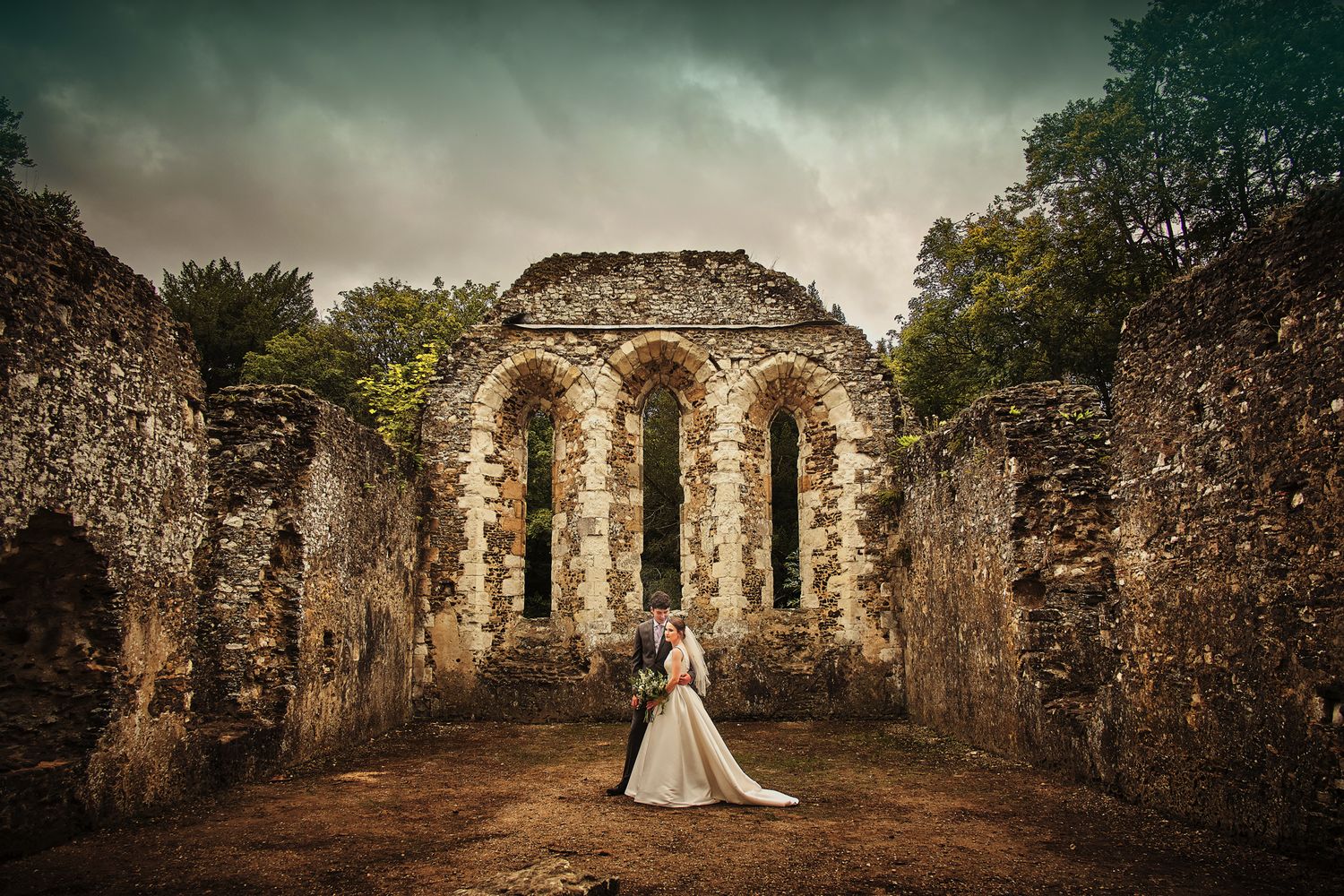 A wedding couple stands beneath dramatic stone arches of an ancient ruined church with moody sky overhead.