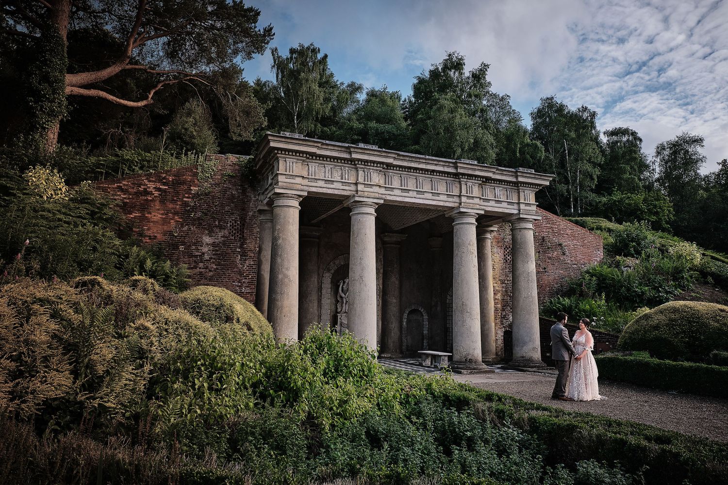 A romantic stone colonnade with classical columns nestled among lush gardens and greenery creates a dreamy architectural setting.
