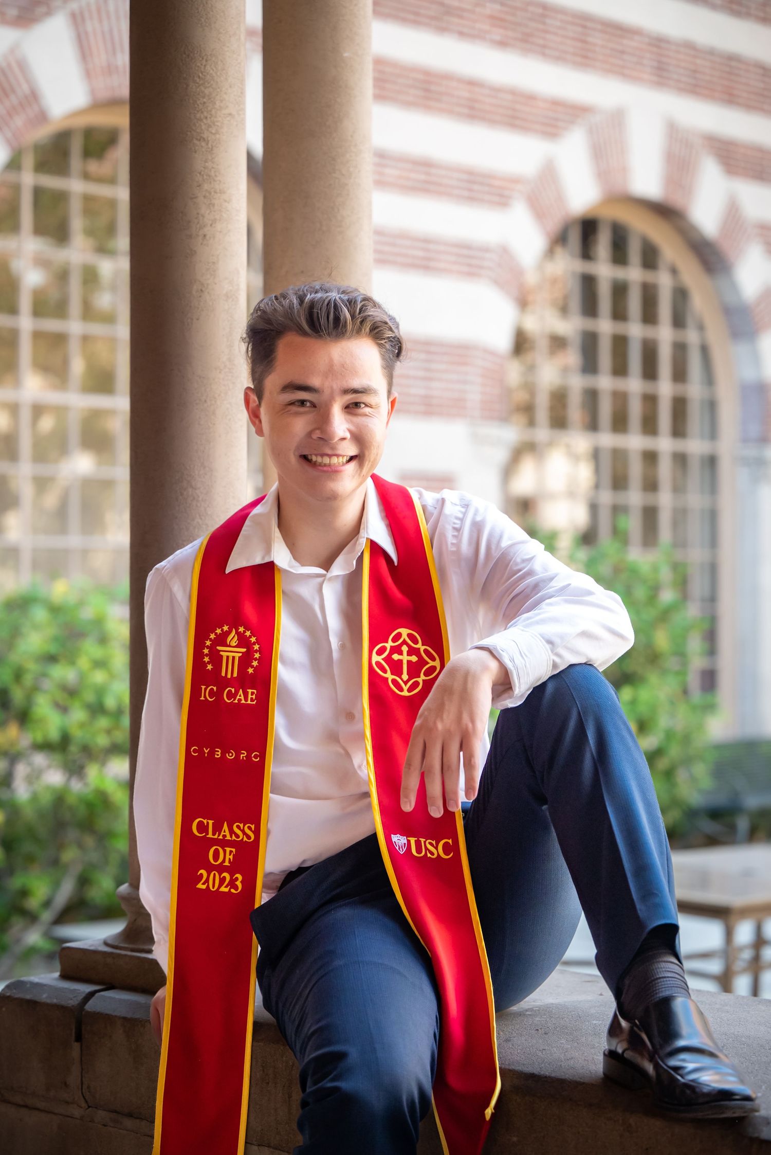 Graduate wearing a red and gold Class of 2023 graduation stole sits casually on a stone ledge at a historic university building.