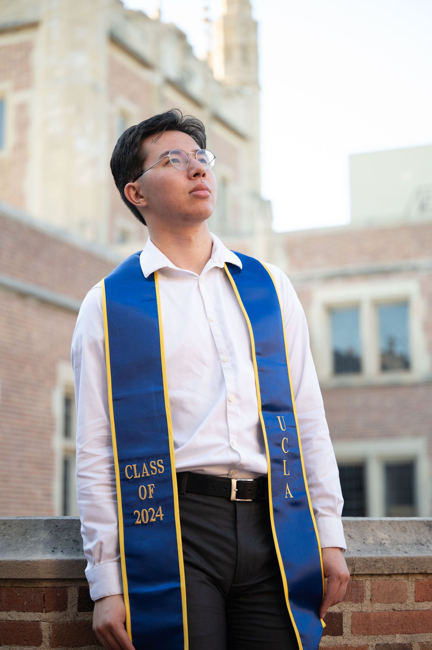 Graduate wearing a blue and gold Class of 2024 stole over white shirt stands against brick wall near historic building.