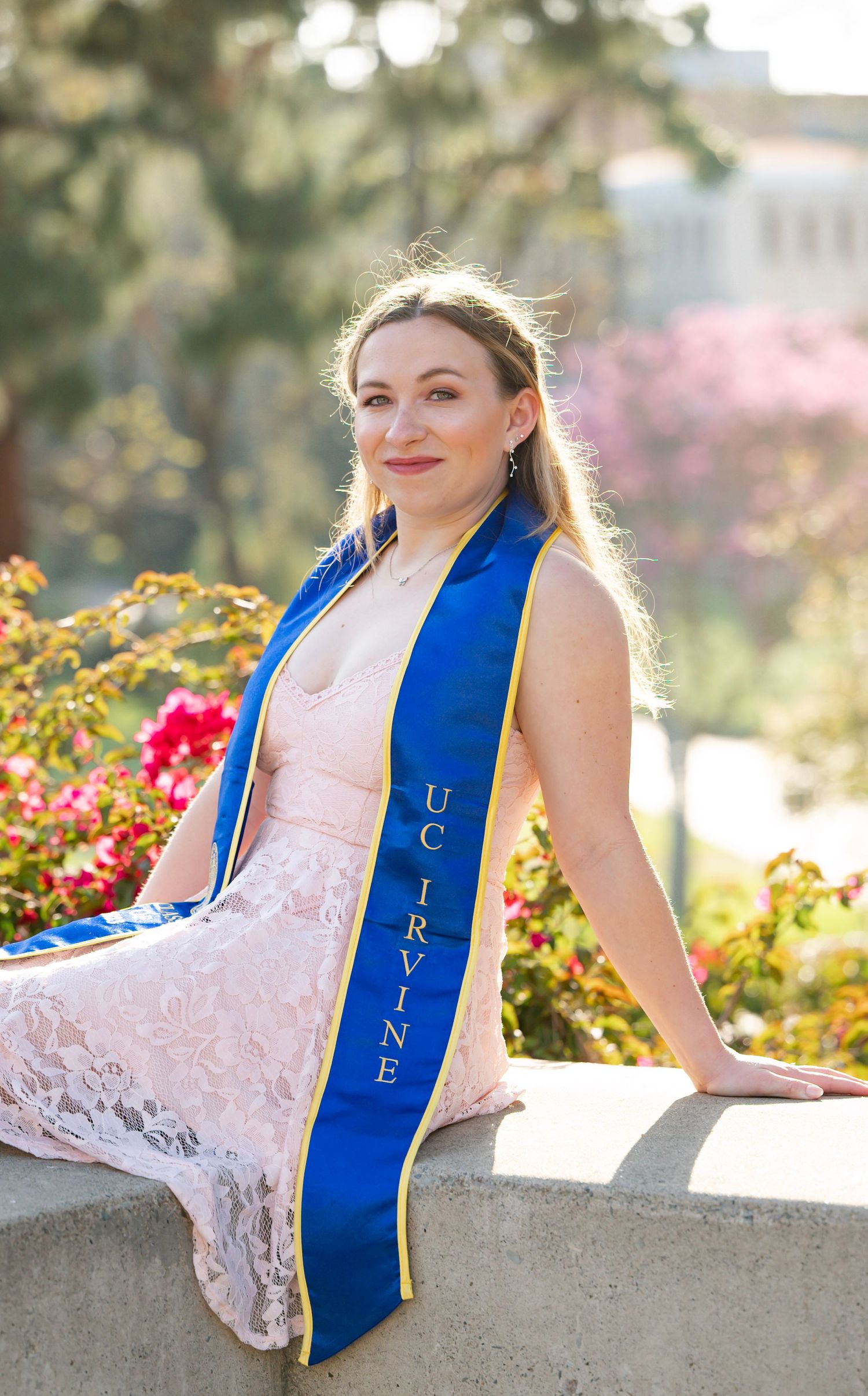 Graduate wearing a pink lace dress and blue UC IRVINE sash poses in garden with blooming flowers in background.