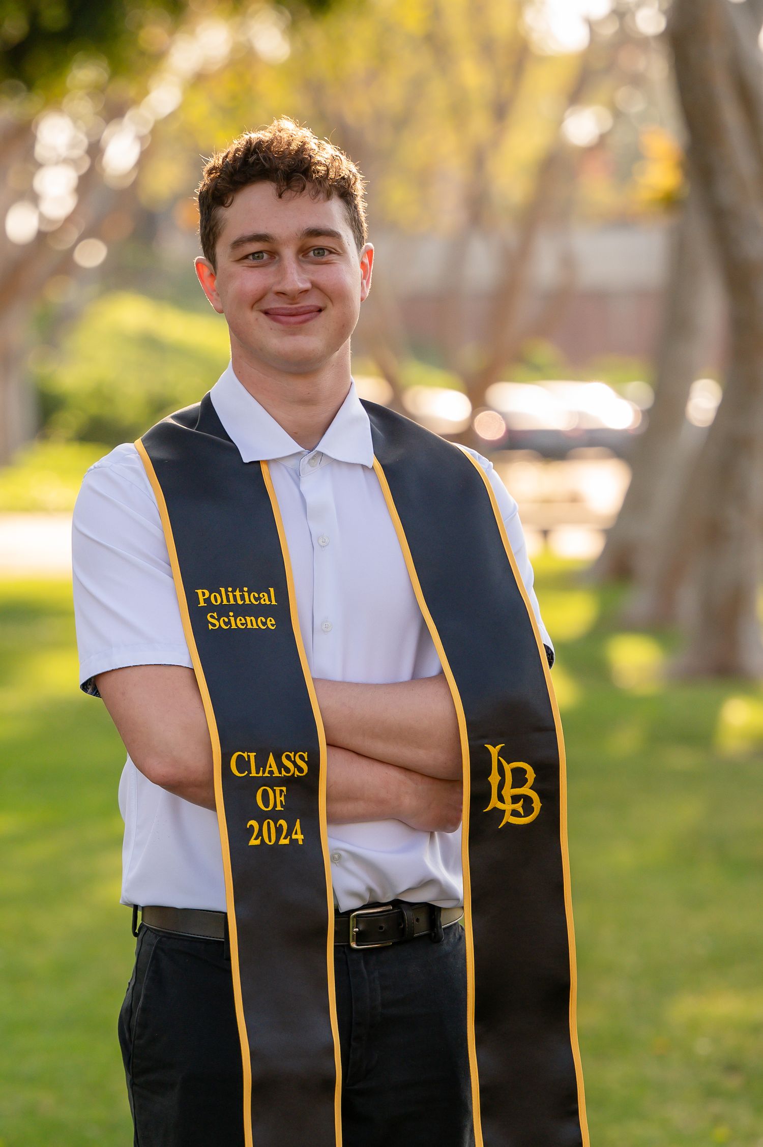 A graduate in formal attire wearing a black and gold stole for Political Science Class of 2024 poses outdoors in sunlit setting.