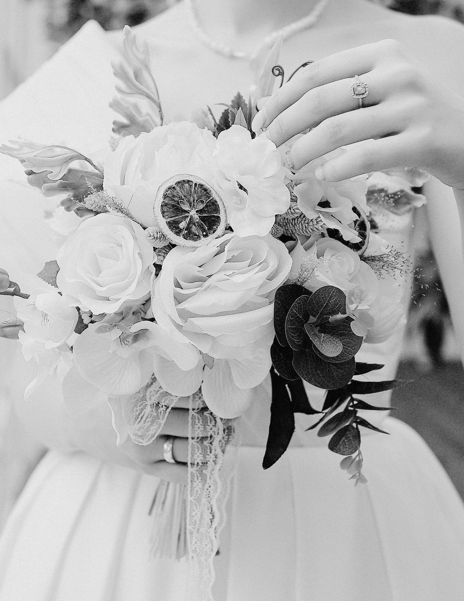 Black and white photo showing a bridal bouquet with white roses and lace details, with a wedding ring visible.