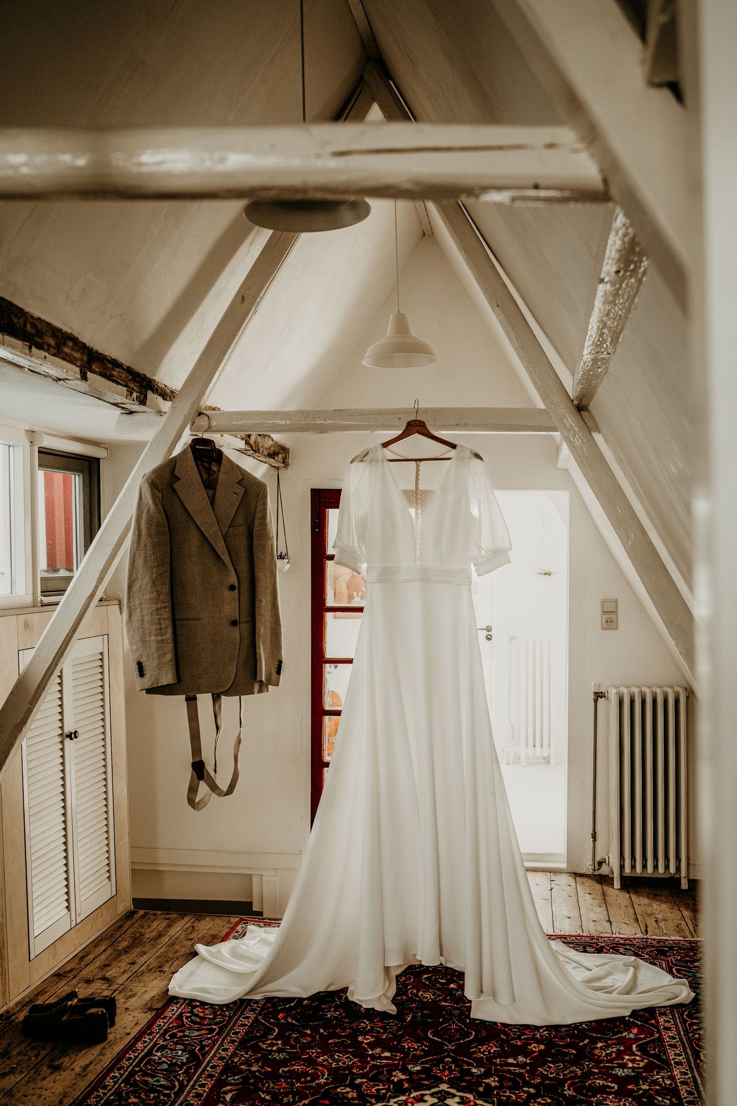 Wedding dress and suit hanging in rustic attic room with exposed beams.