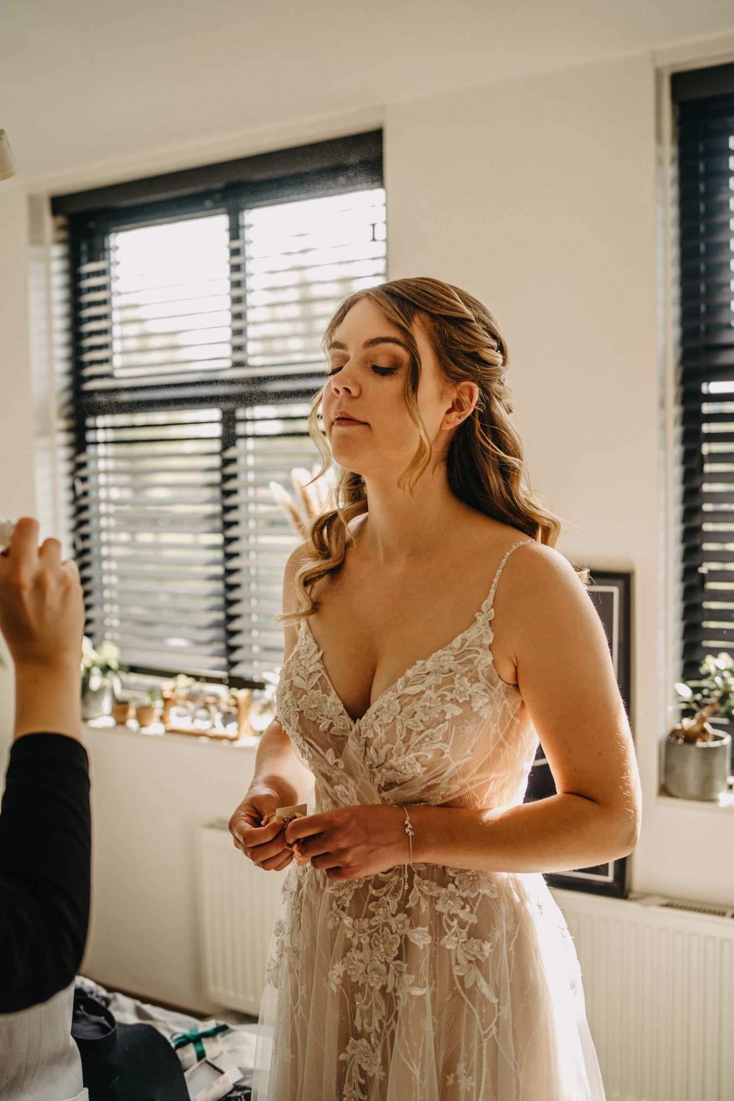 A delicate lace wedding dress being fitted in a naturally lit room with window blinds.