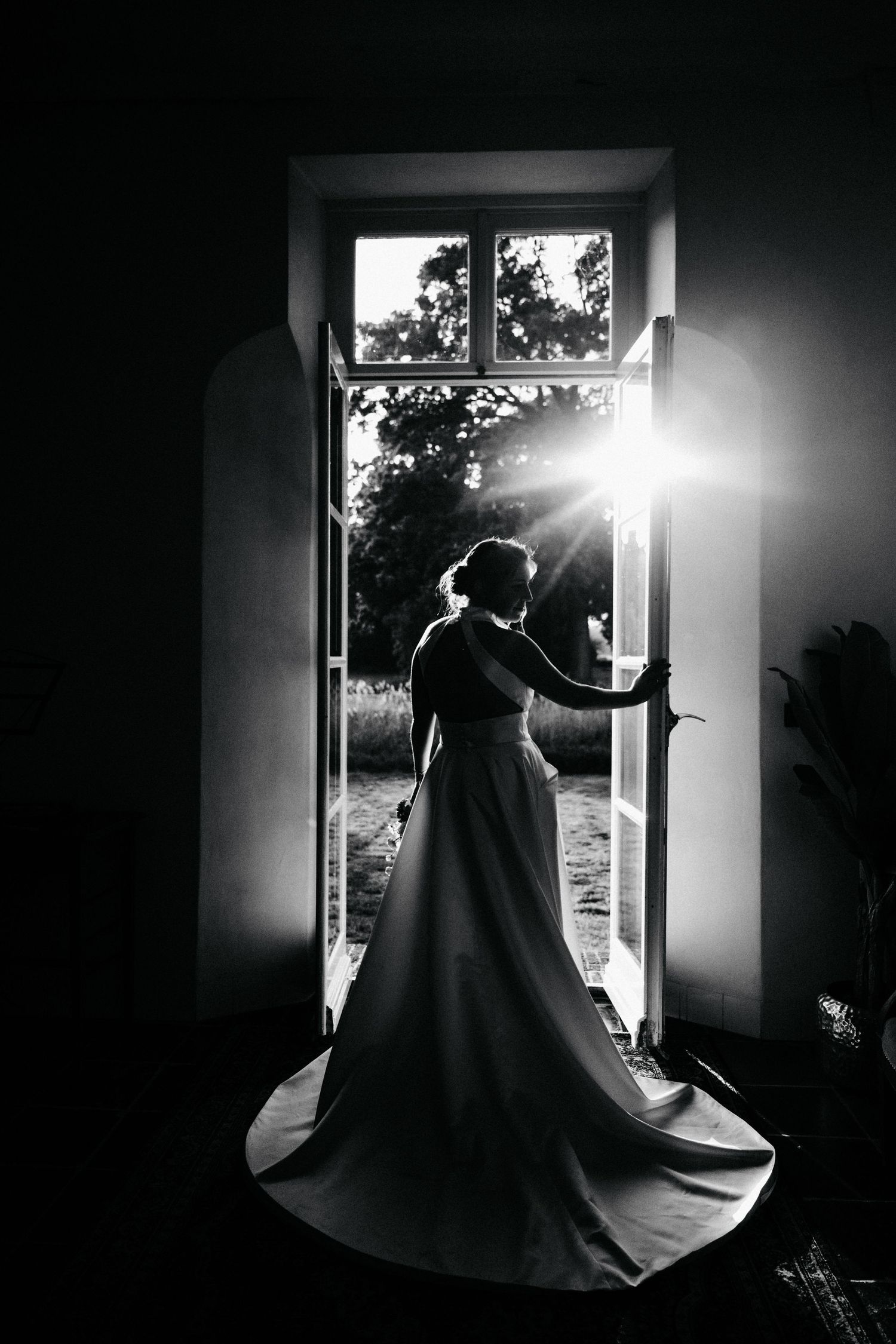 A silhouette of a bride in a flowing dress standing in a sunlit doorway.