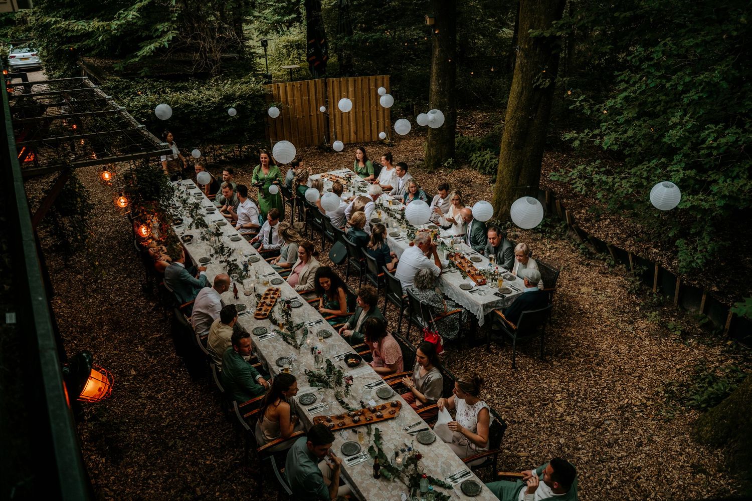 An outdoor wedding reception dinner setup in a forest with white decor and string lights.