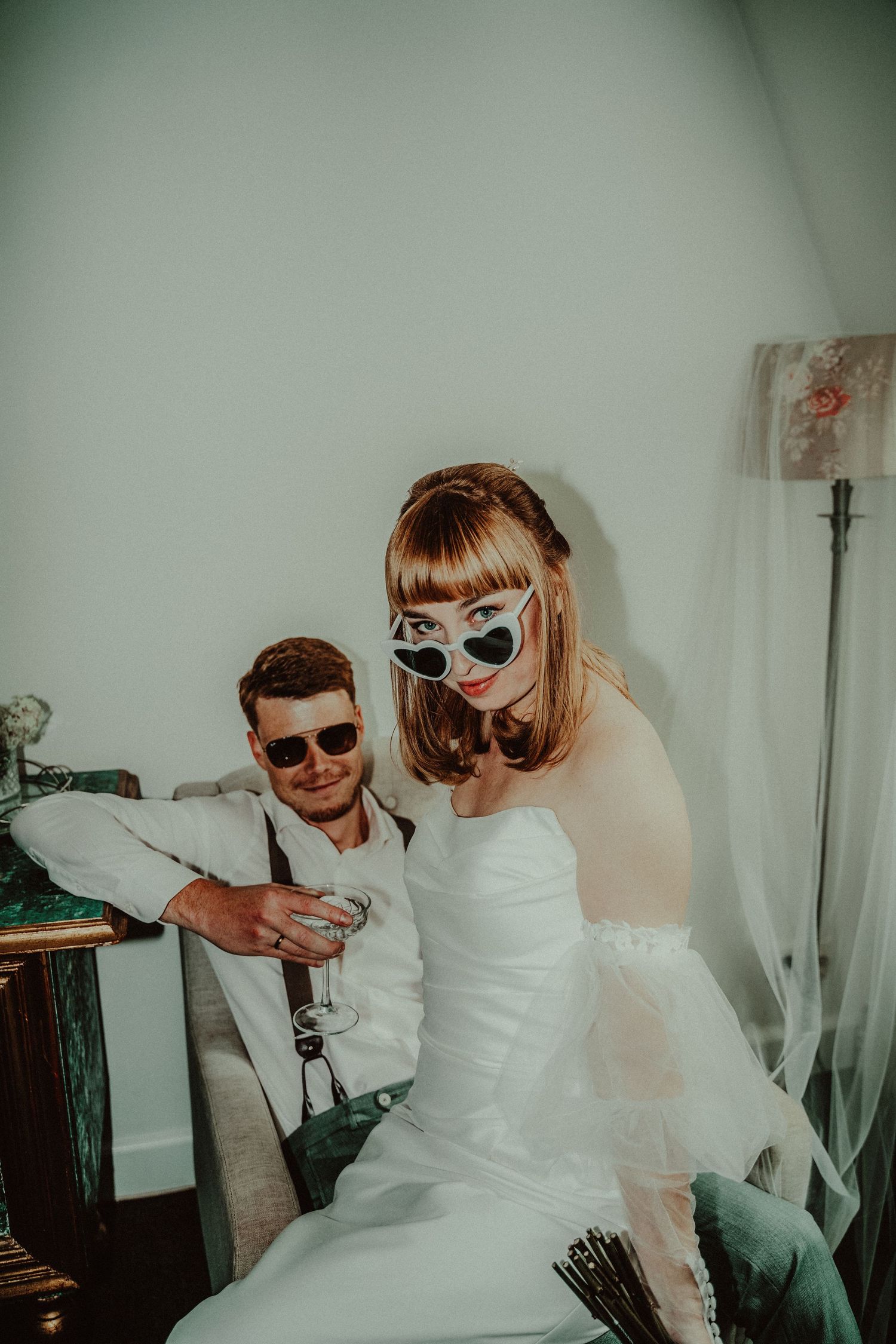 A bride in sunglasses and white dress poses with someone in suspenders during a wedding photoshoot.