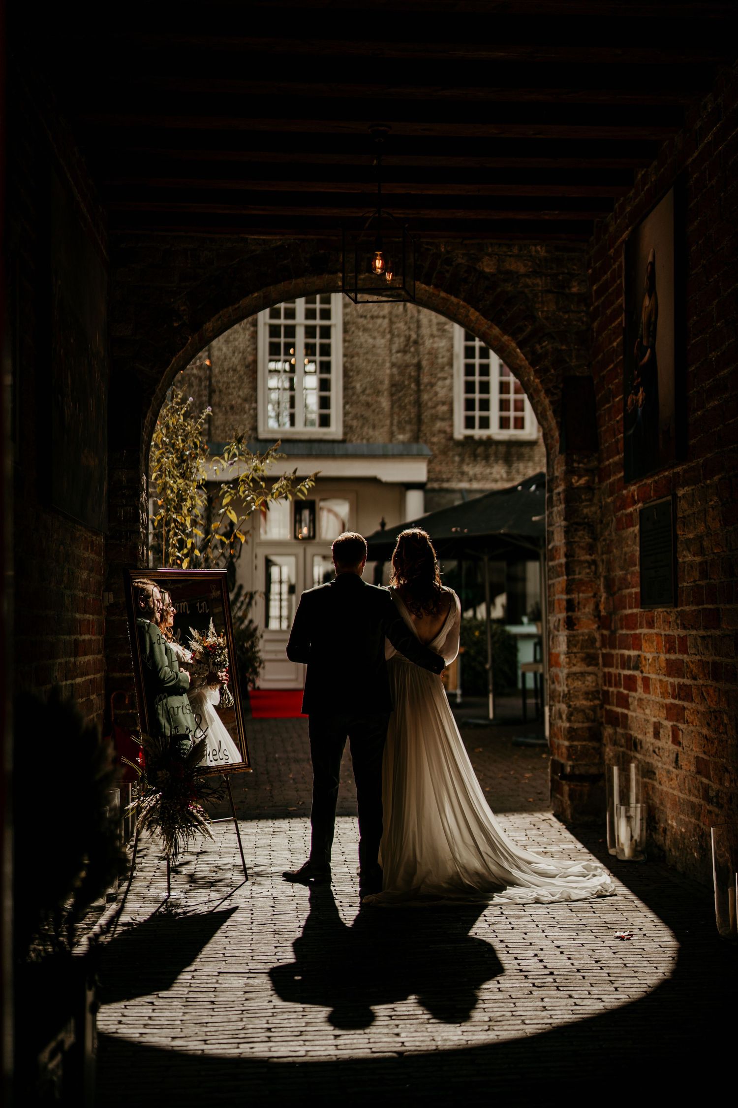 Wedding couple stands in silhouette under historic stone archway with dramatic lighting.