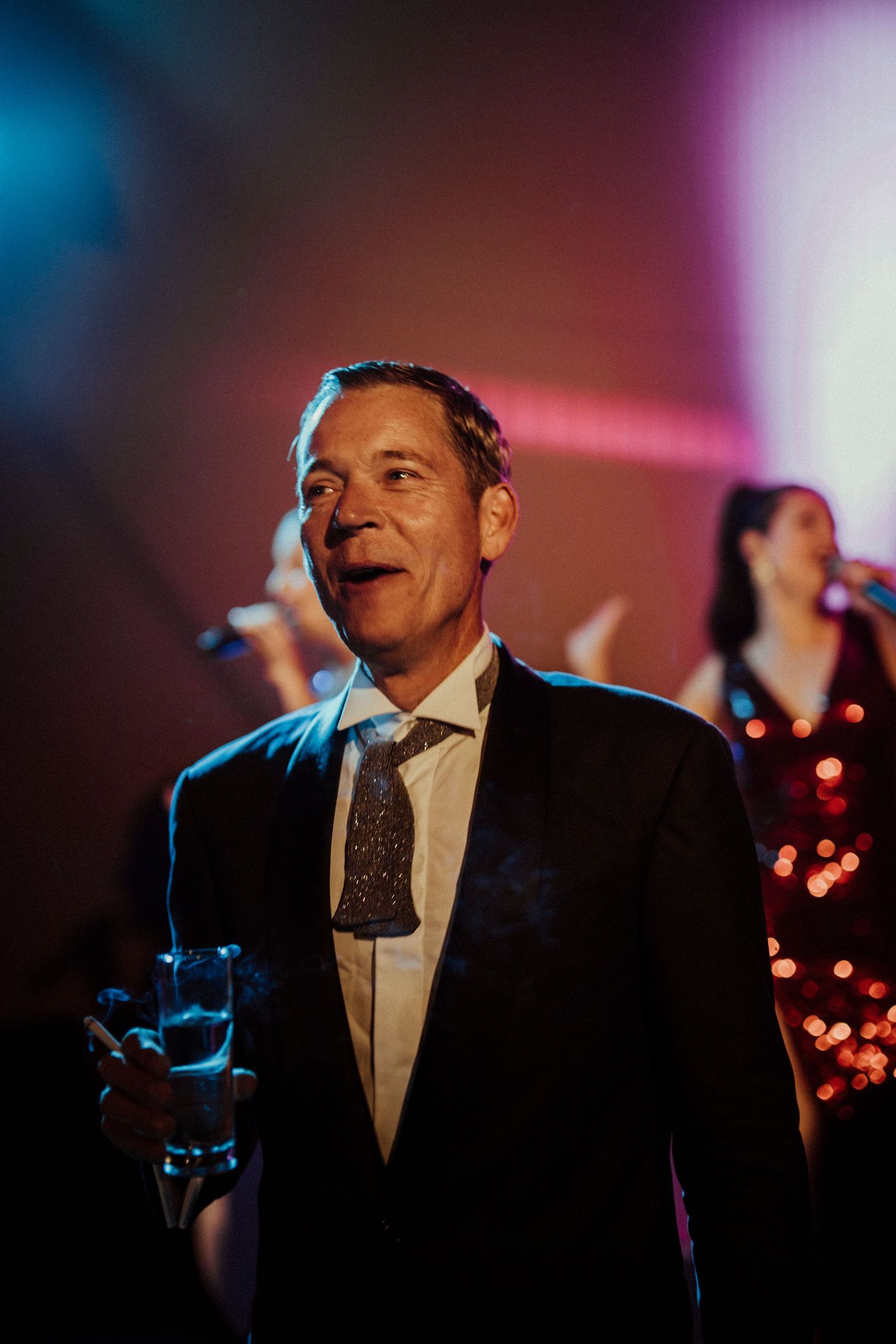 A performer in a black suit and tie sings on stage with colorful stage lighting and decorative lights in the background.