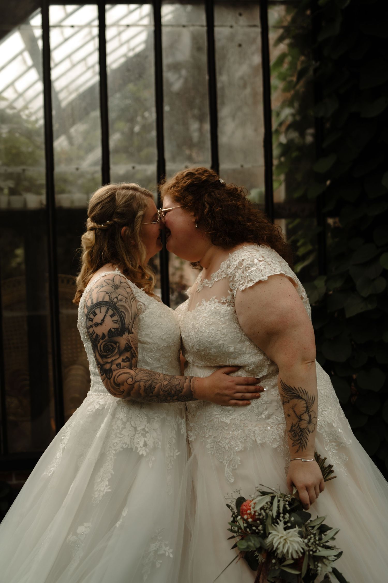 Two brides in white lace wedding dresses embrace in front of greenhouse windows.