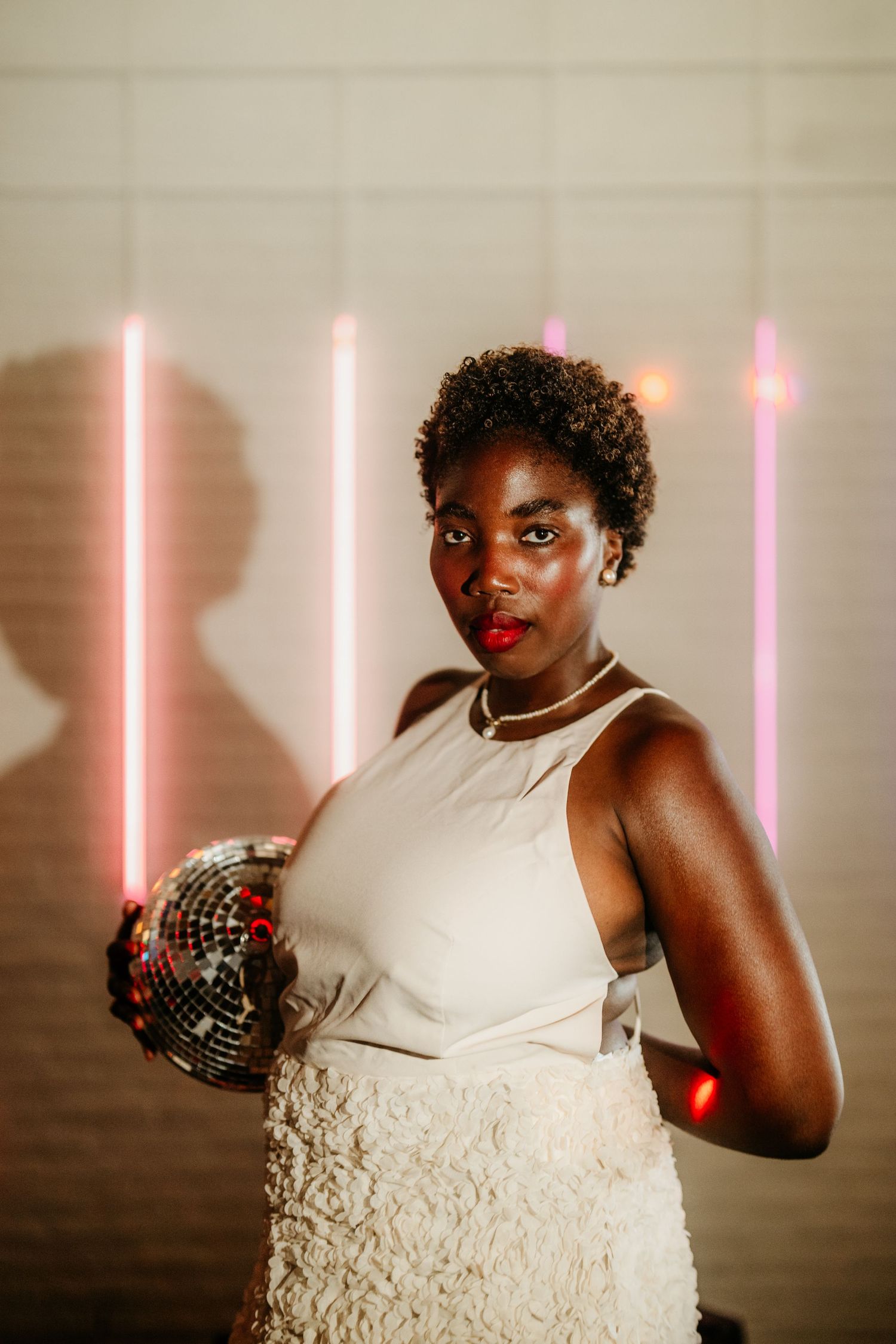 A figure in a cream-colored dress holds a disco ball against a backdrop of pink neon lights.