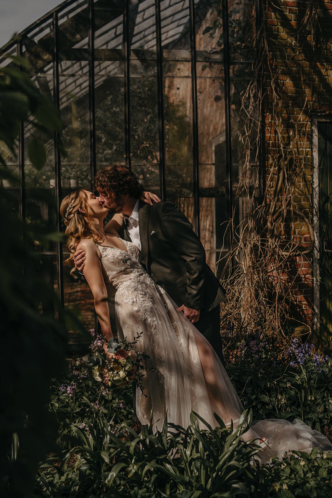Romantic wedding portrait against greenhouse with trailing vines and natural lighting.