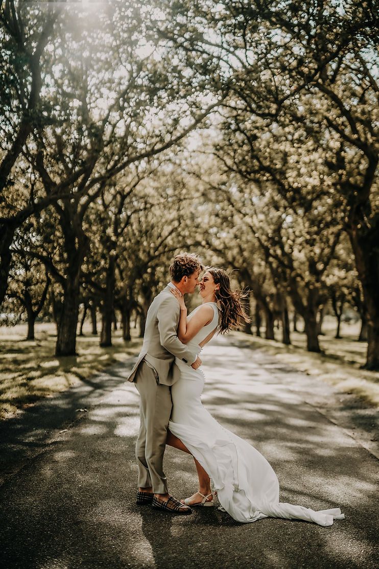 Newlyweds embrace in a sunlit tree-lined avenue wearing elegant wedding attire.