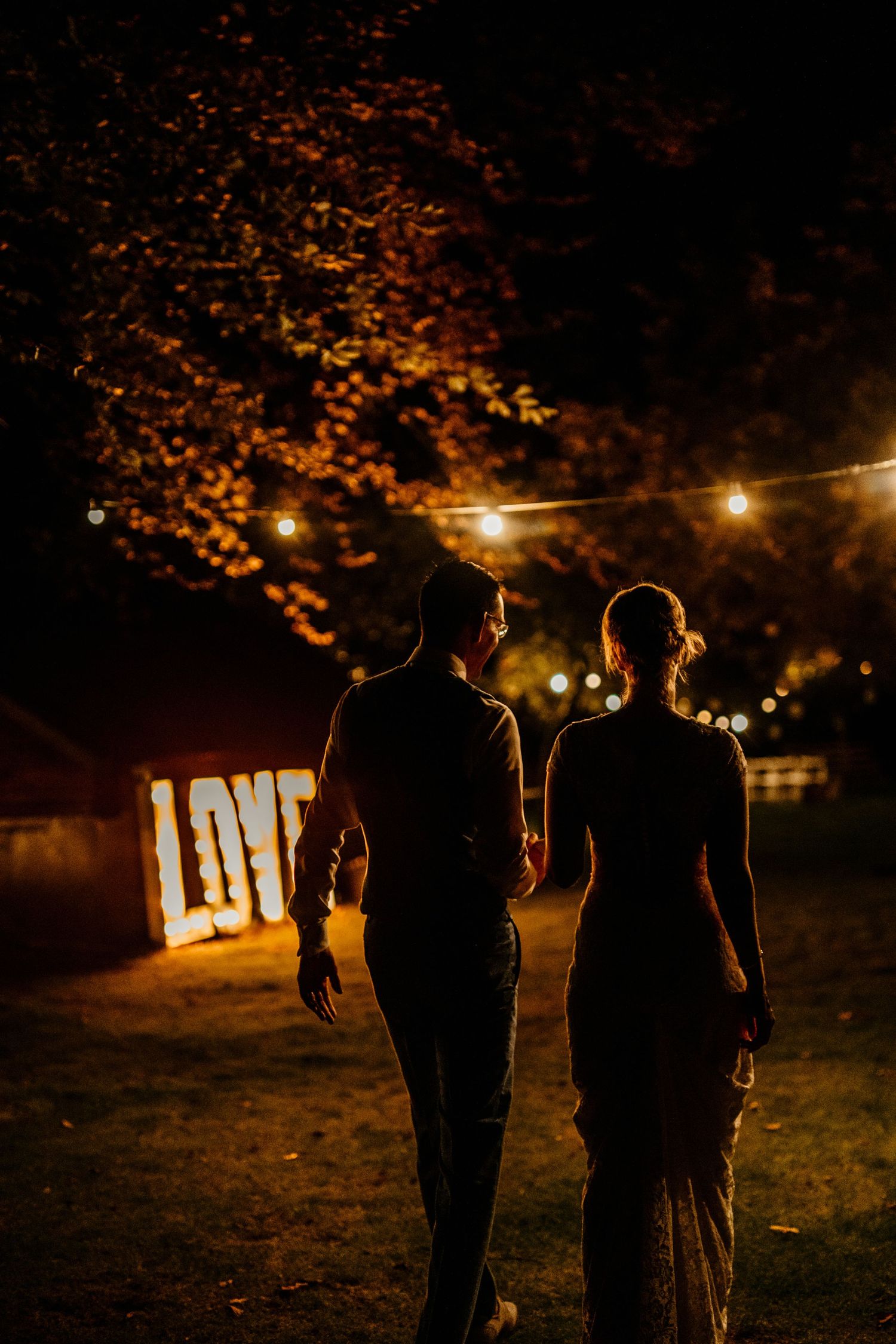 Couple walks under string lights at night with illuminated LOVE sign in background.