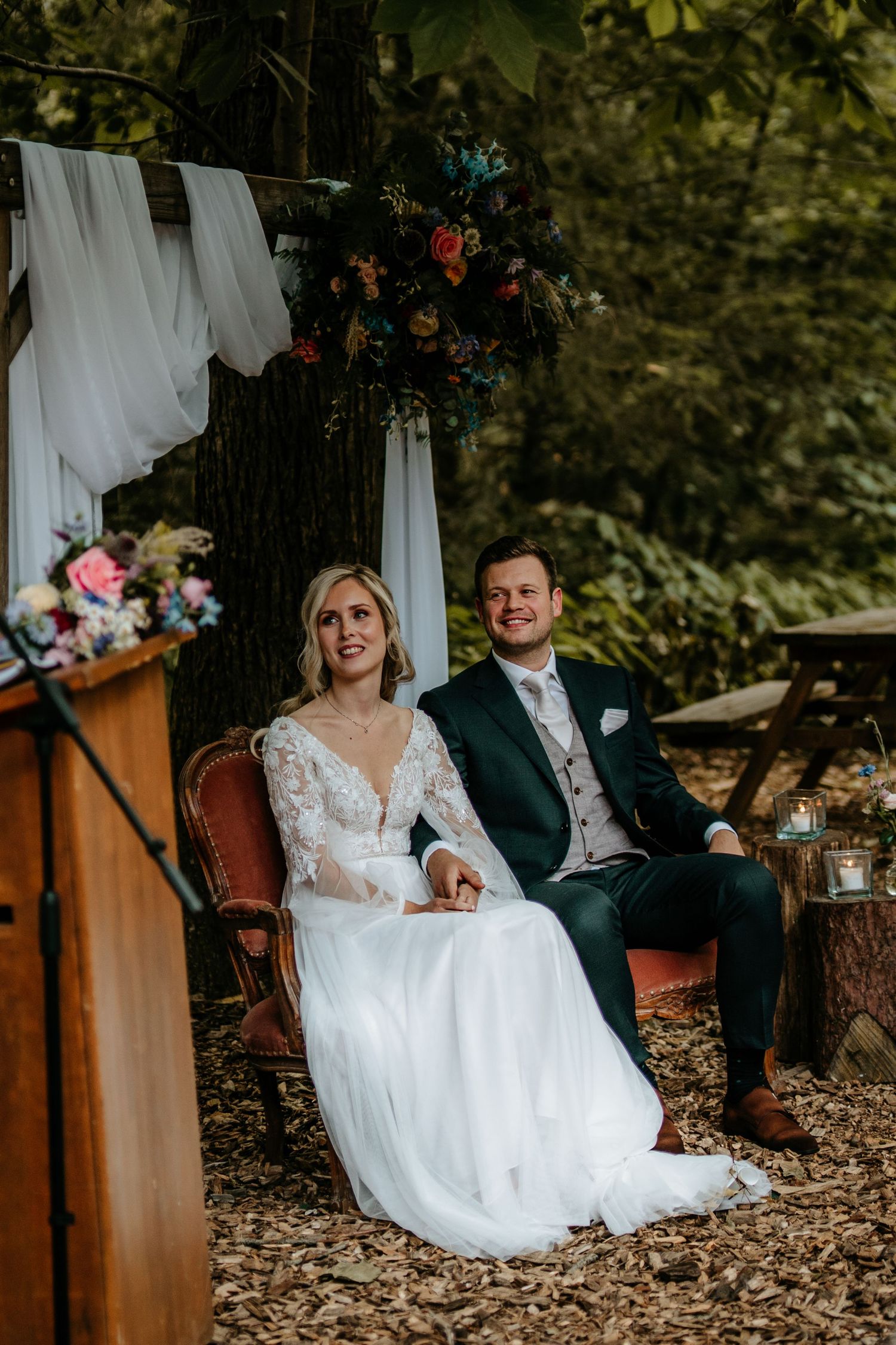 A couple sits in ornate chairs during their woodland wedding ceremony.