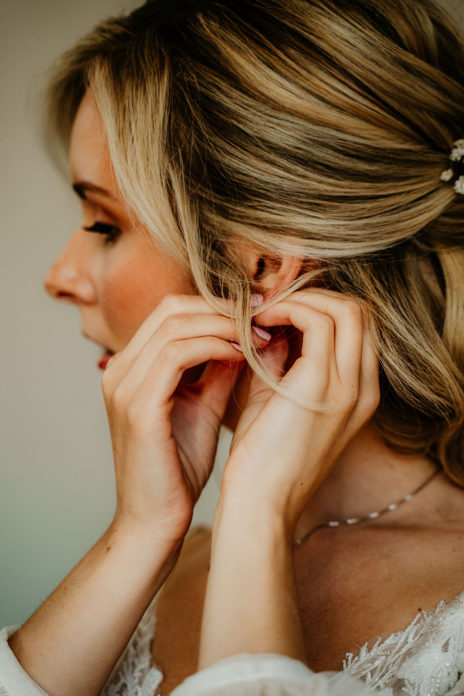 Close-up profile shot of a blonde woman putting on jewelry while wearing a white lace gown.