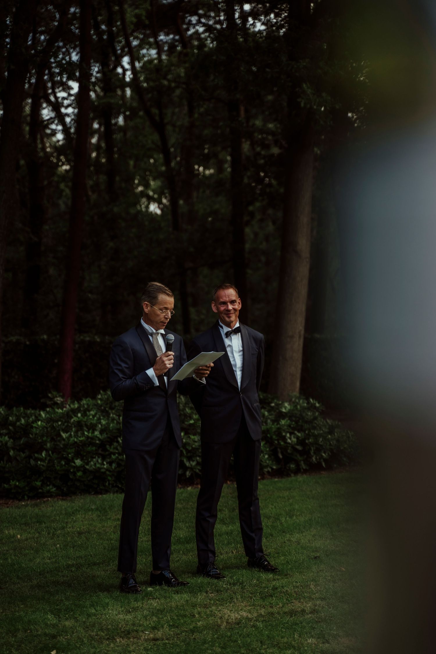 Two men in tuxedos share a moment while reading documents in a garden setting.