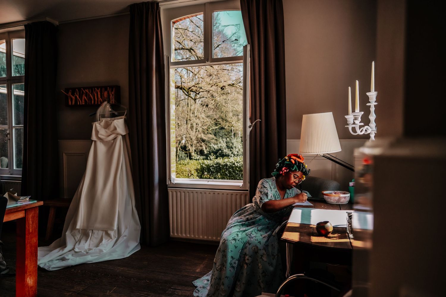 Elegant wedding gown hangs by window in moody brown room with upholstered chair and antique lamp.