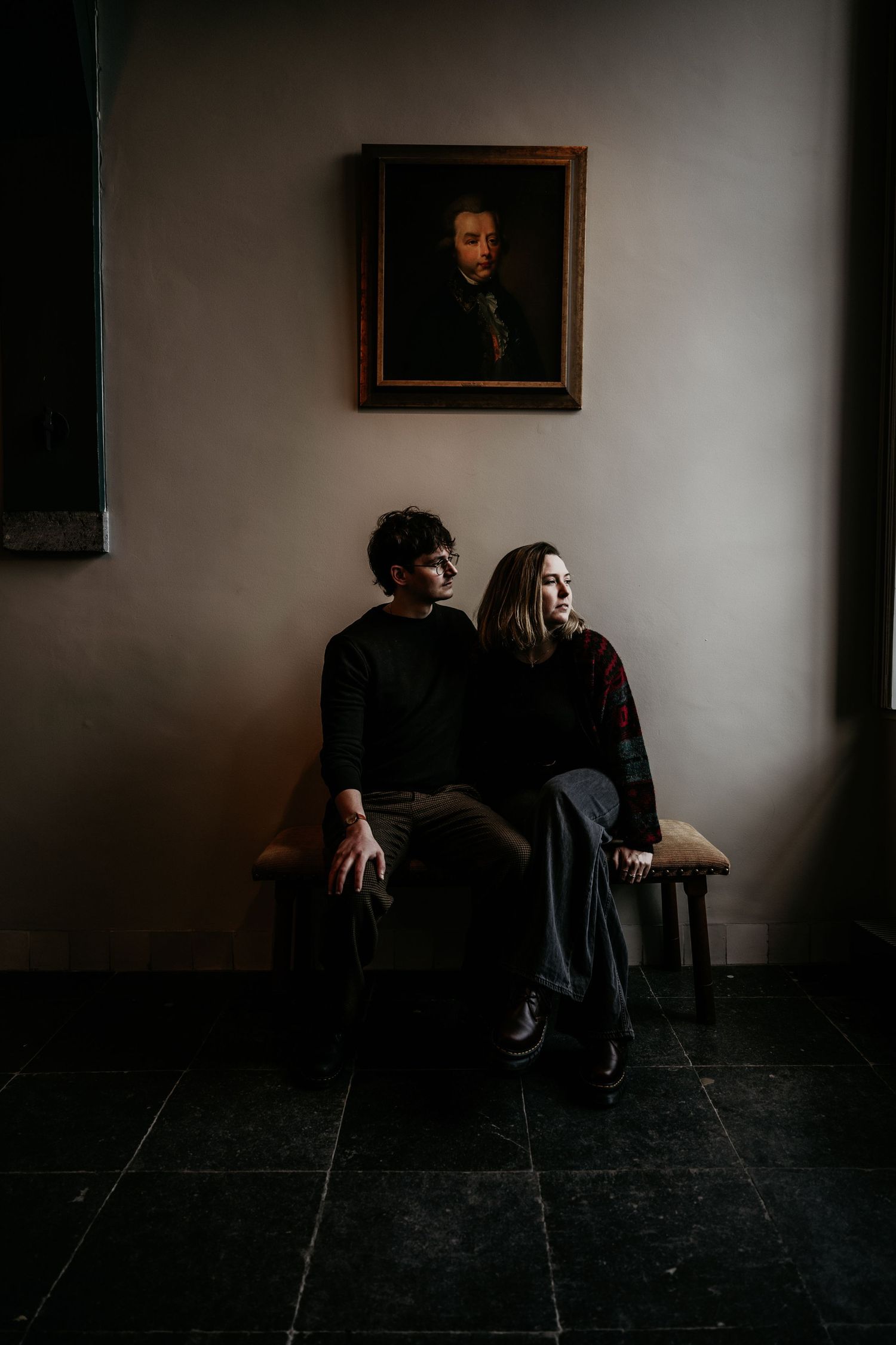 Two people in dark clothing sit together on a bench beneath an old portrait in a moody, dimly lit interior setting.