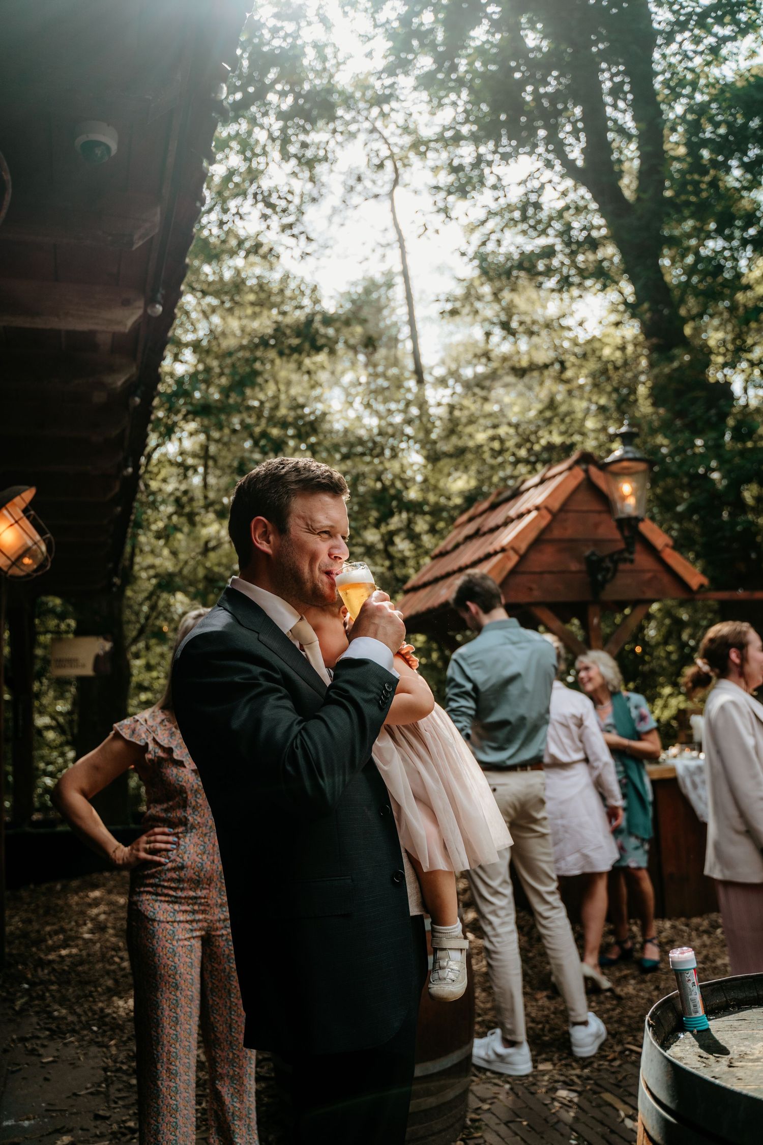 Wedding guest enjoys drink at outdoor reception under forest canopy.